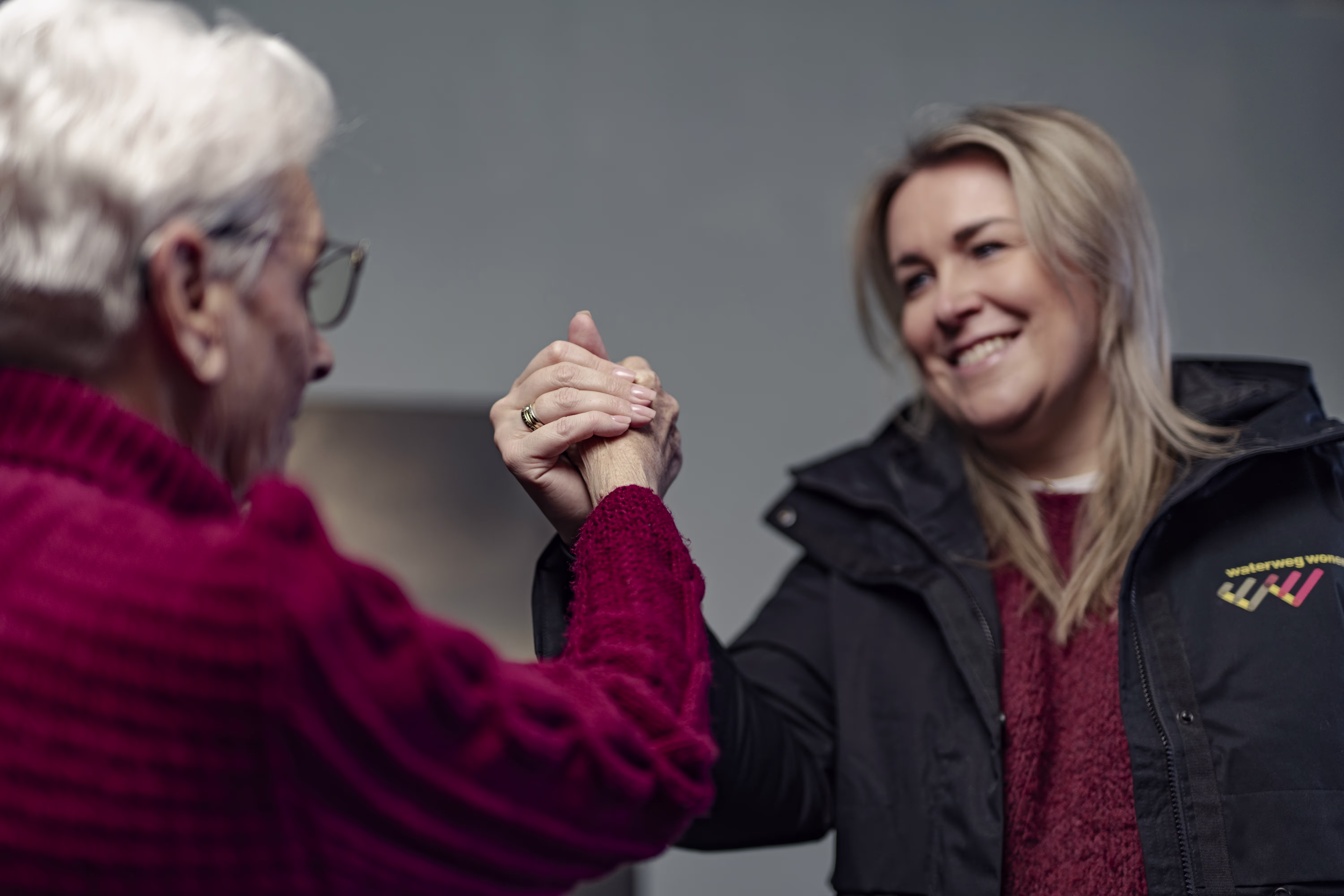 An older woman and a younger blonde woman shake hands and smile at each other; both are wearing red sweaters.