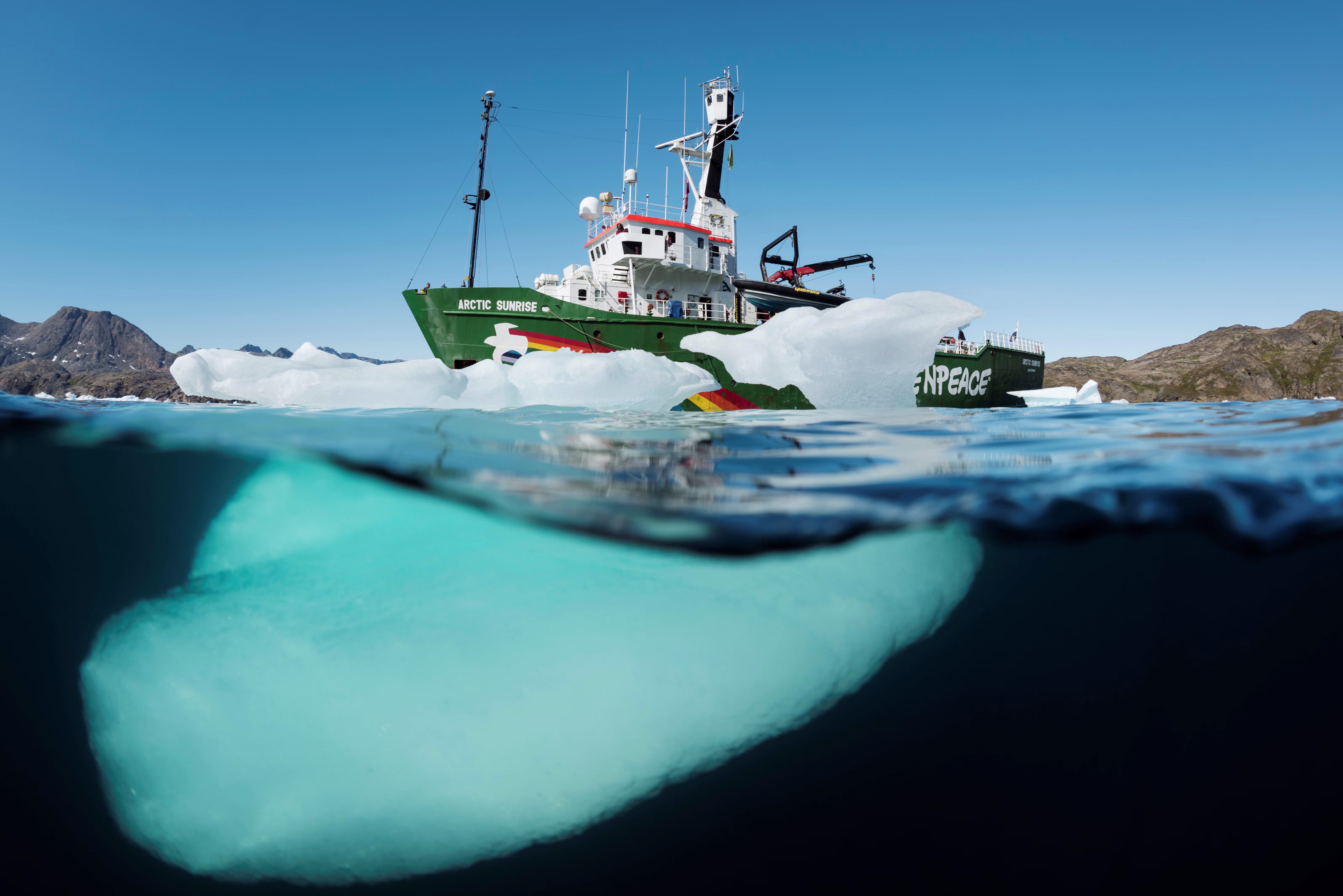Een Greenpeace-schip vaart tussen ijsschotsen in arctische wateren, gefotografeerd vanuit het water met ijsberg op voorgrond.