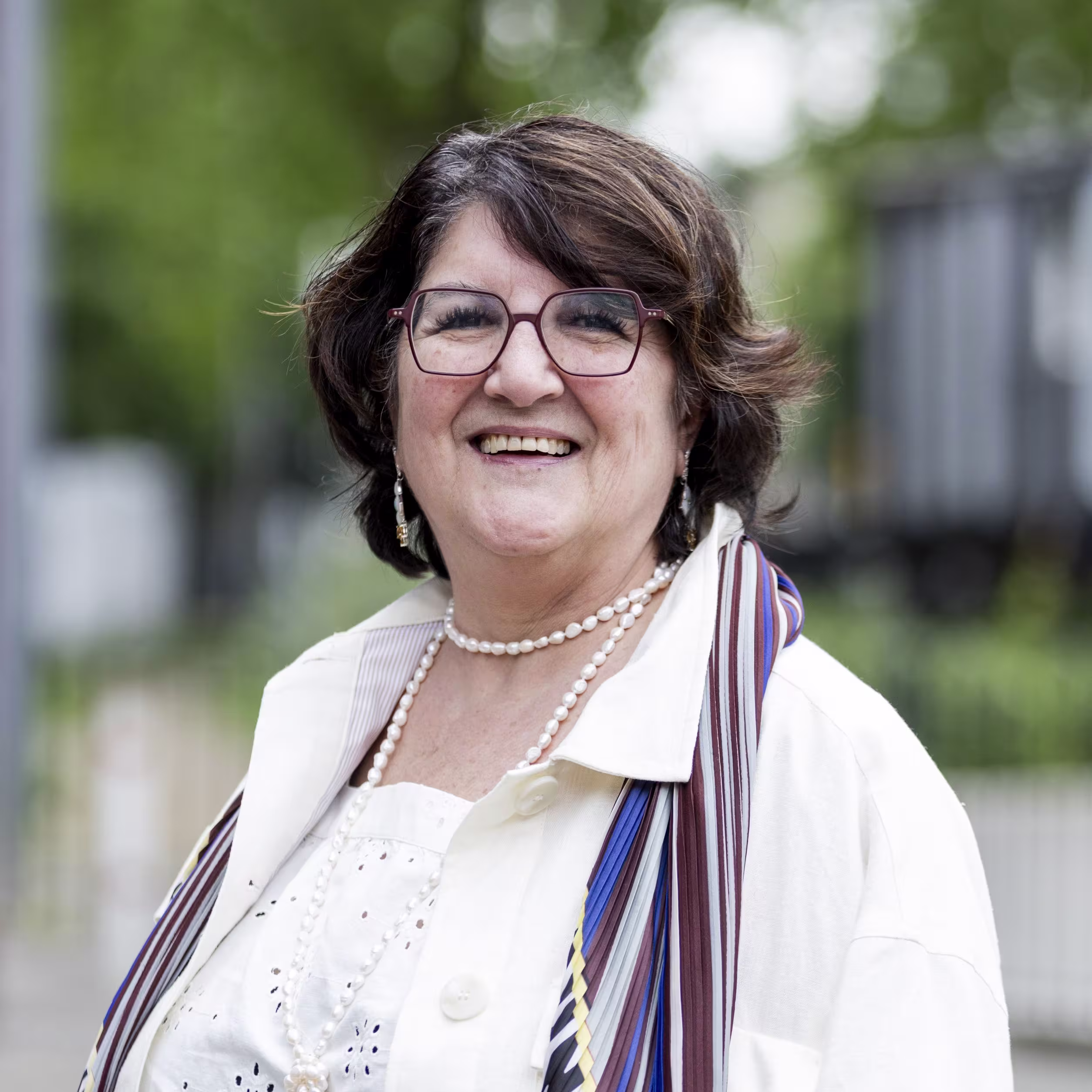 Lachende vrouw met bril, parelketting en kleurrijke sjaal poseert buiten voor een professionele foto.