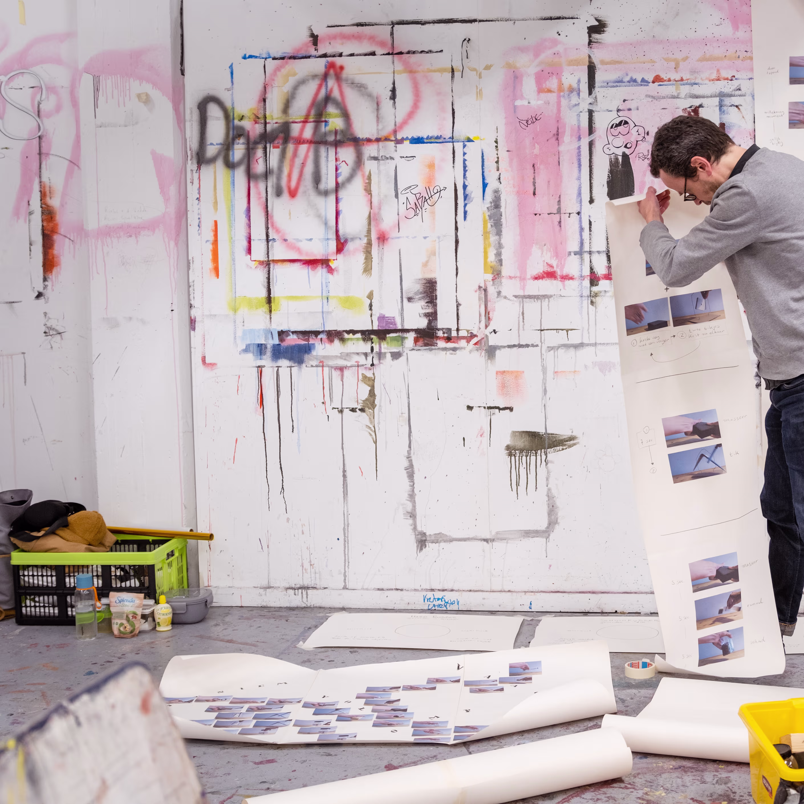 Een kunstenaar werkt in zijn atelier met verf en foto's, omringd door kleurrijke schilderijen aan de muur.