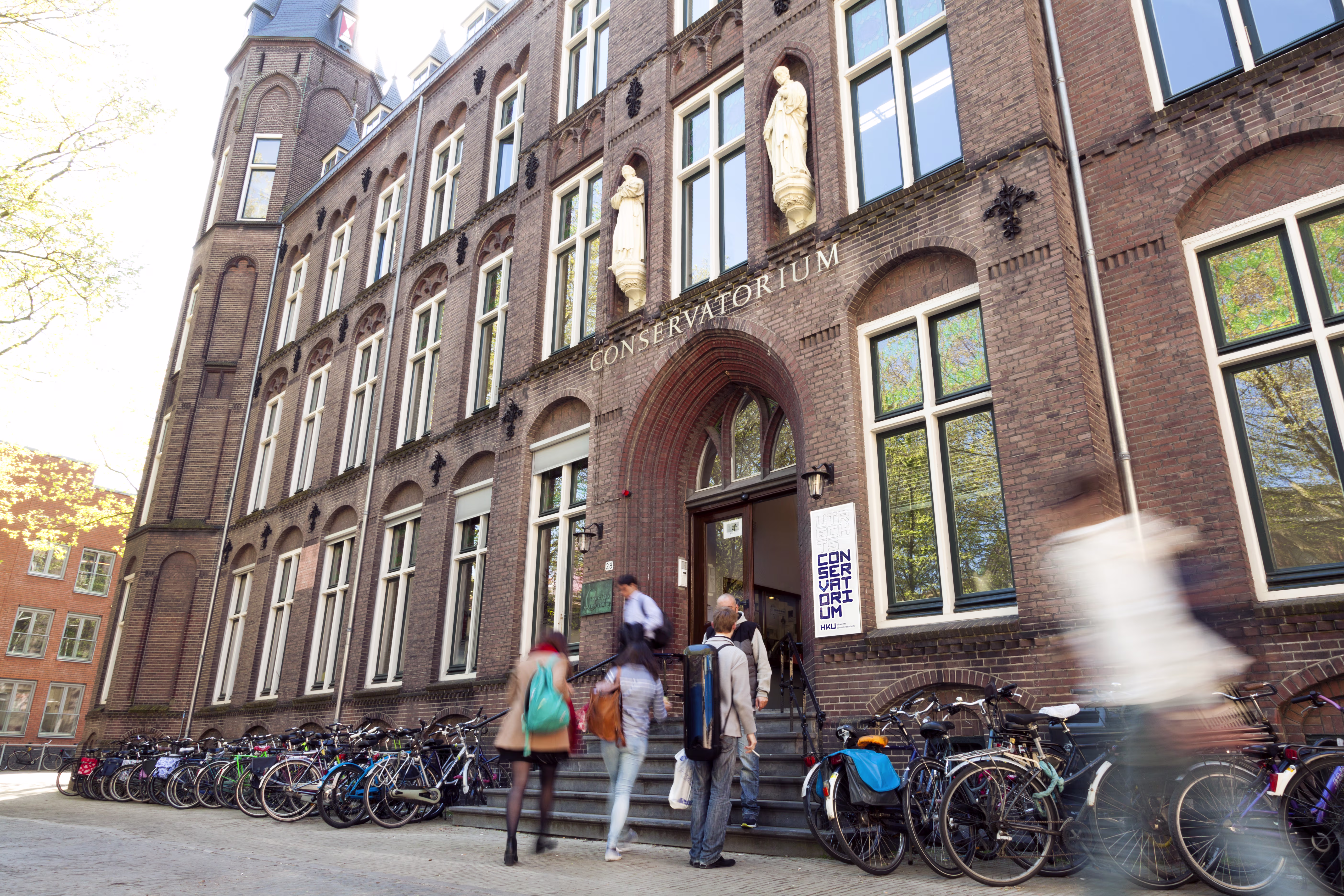 Studenten lopen de trap op naar het Conservatorium gebouw van HKU, met veel fietsen geparkeerd voor de ingang.