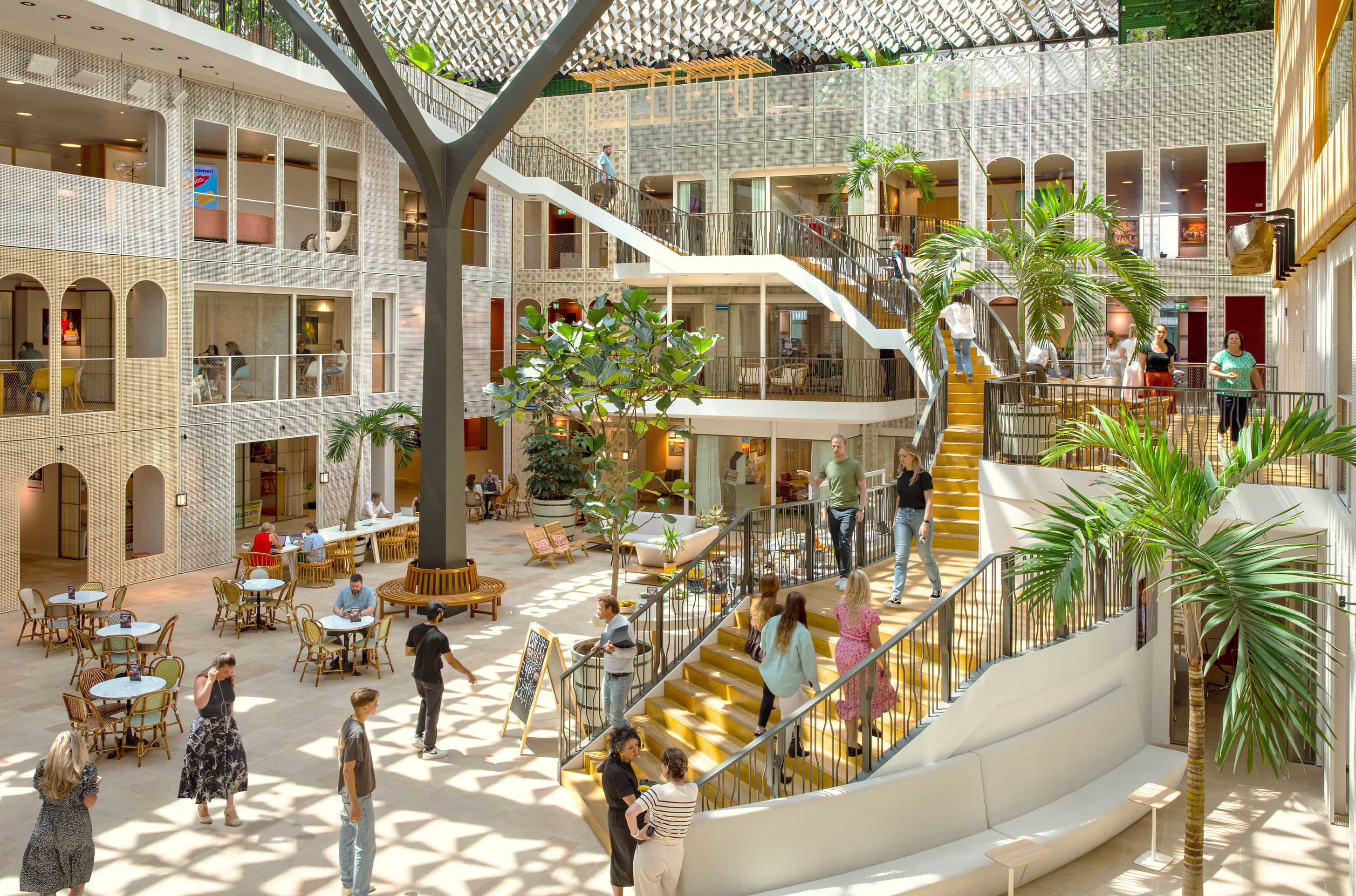 Moderne kantooromgeving met atrium, trappen en groenvoorziening waar mensen werken en ontmoeten in een open architectuur.