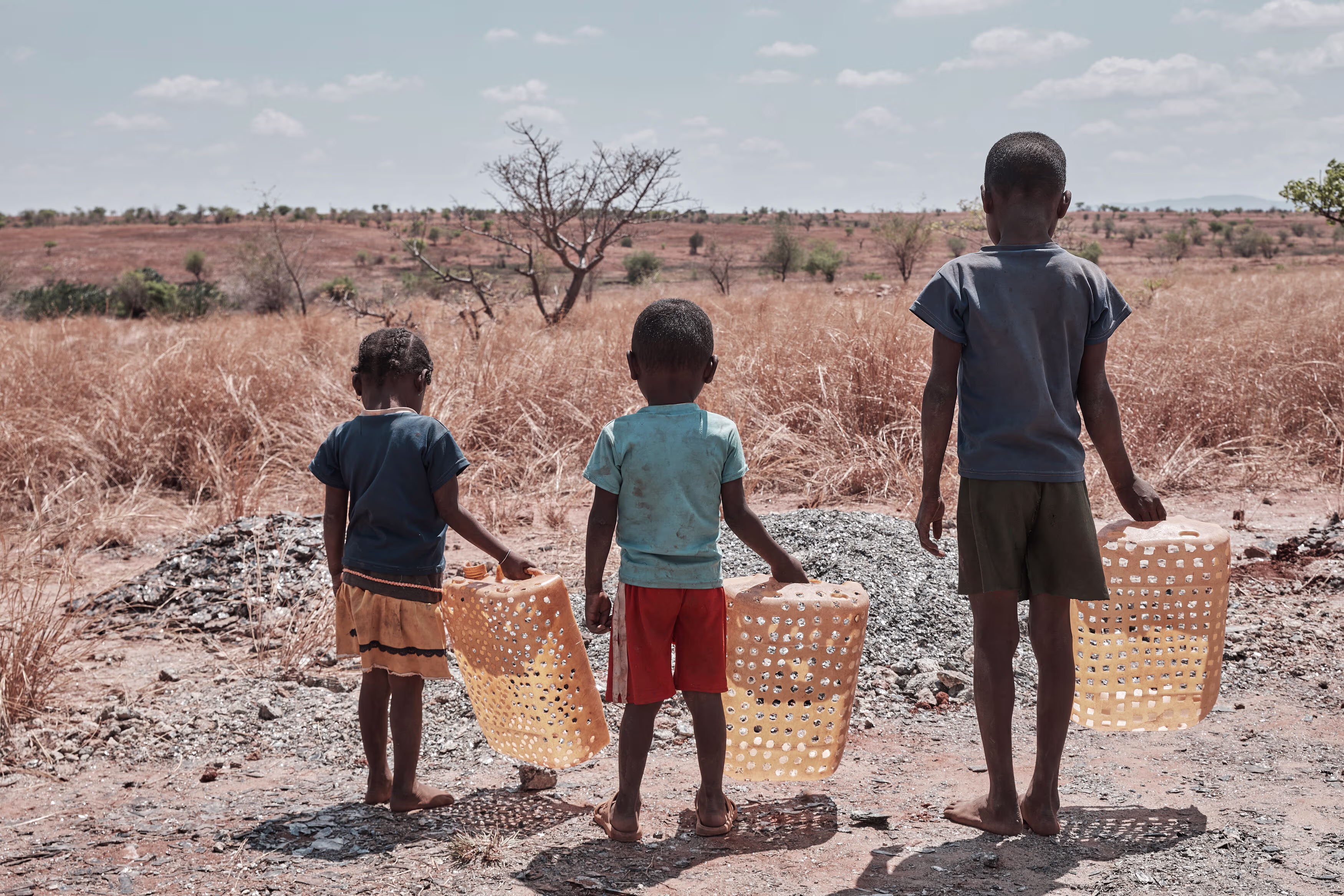Drie kinderen met plastic kratten lopen door een droog, kaal landschap met verspreide bomen en struiken.