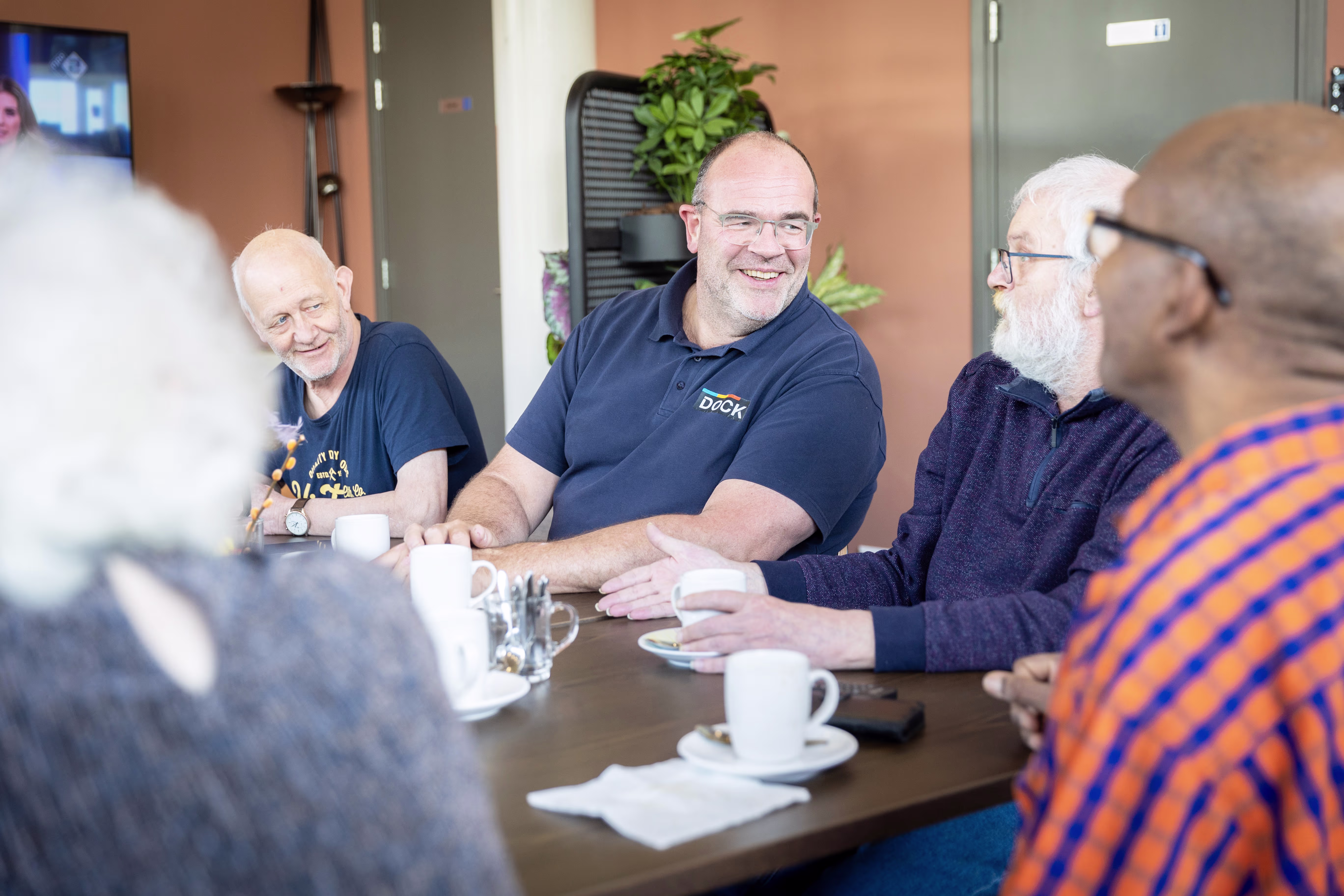 Een groep mannen zit lachend rond een tafel met koffiekopjes in een moderne kantooromgeving tijdens een informeel gesprek.