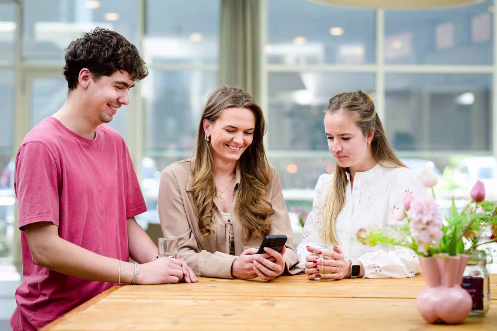 Drie collega's bekijken samen een smartphone aan een houten tafel in een lichte, moderne kantoorruimte.