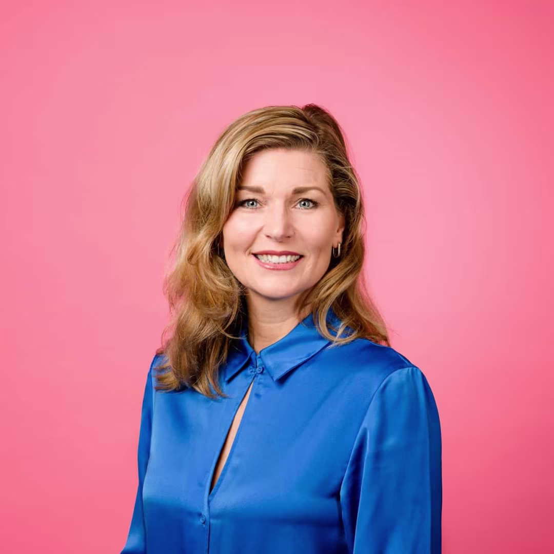 Lachende vrouw met blond haar in blauw satijnen blouse, gefotografeerd tegen een roze achtergrond.