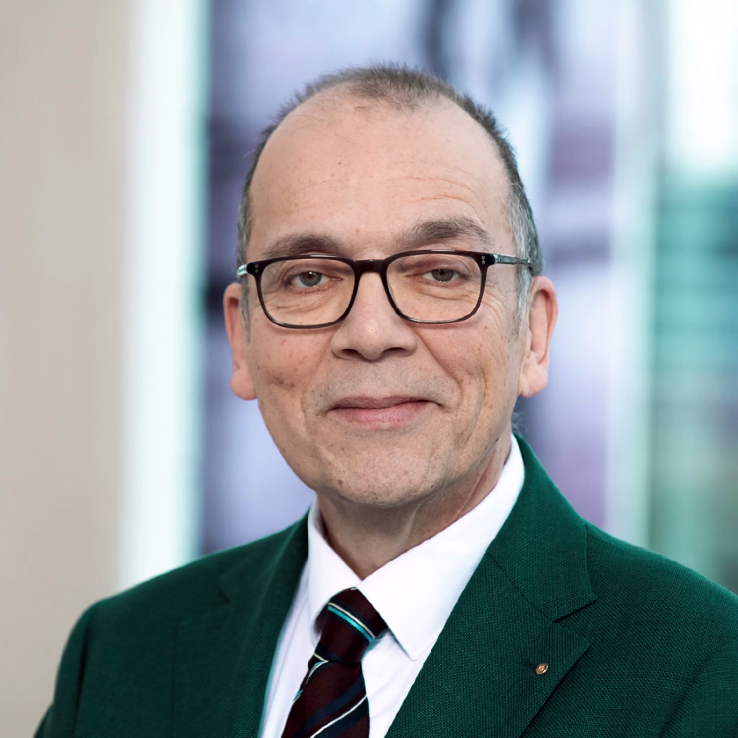 Portrait photo of a man wearing glasses, a green suit, and a striped tie, smiling against a light background.