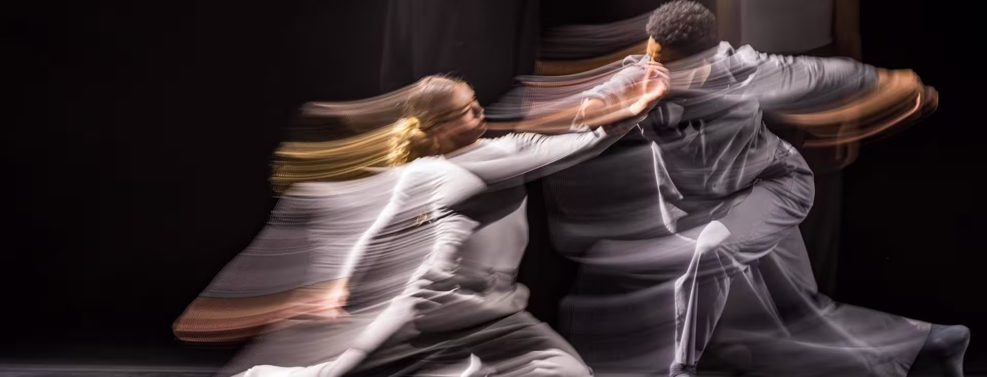 Two dancers in gray outfits move dynamically together, captured with motion blur against a dark background.