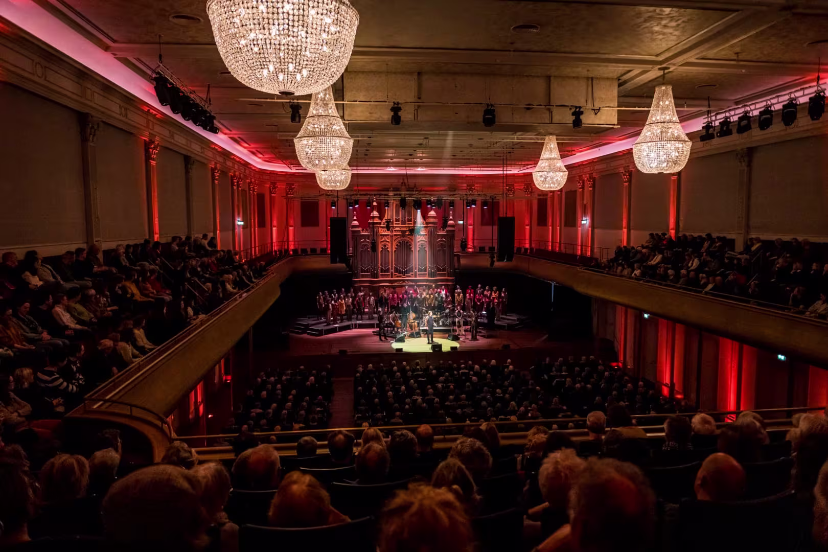 A packed concert hall with red lighting, chandeliers, and a choir on stage in front of a large organ.