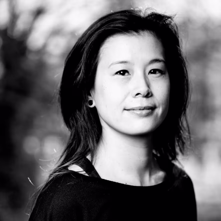 Portretfoto van een jonge vrouw met donker haar, glimlachend, in zwart-wit, buiten met wazige achtergrond.