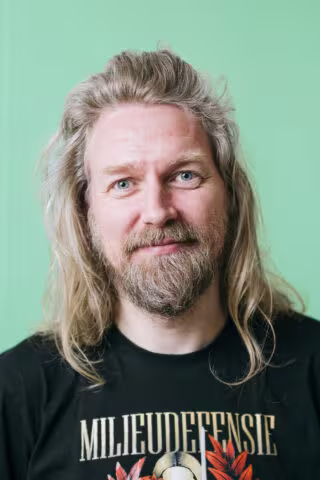 A man with long blond hair and a beard is wearing a black Milieudefensie T-shirt, standing in front of a green background.