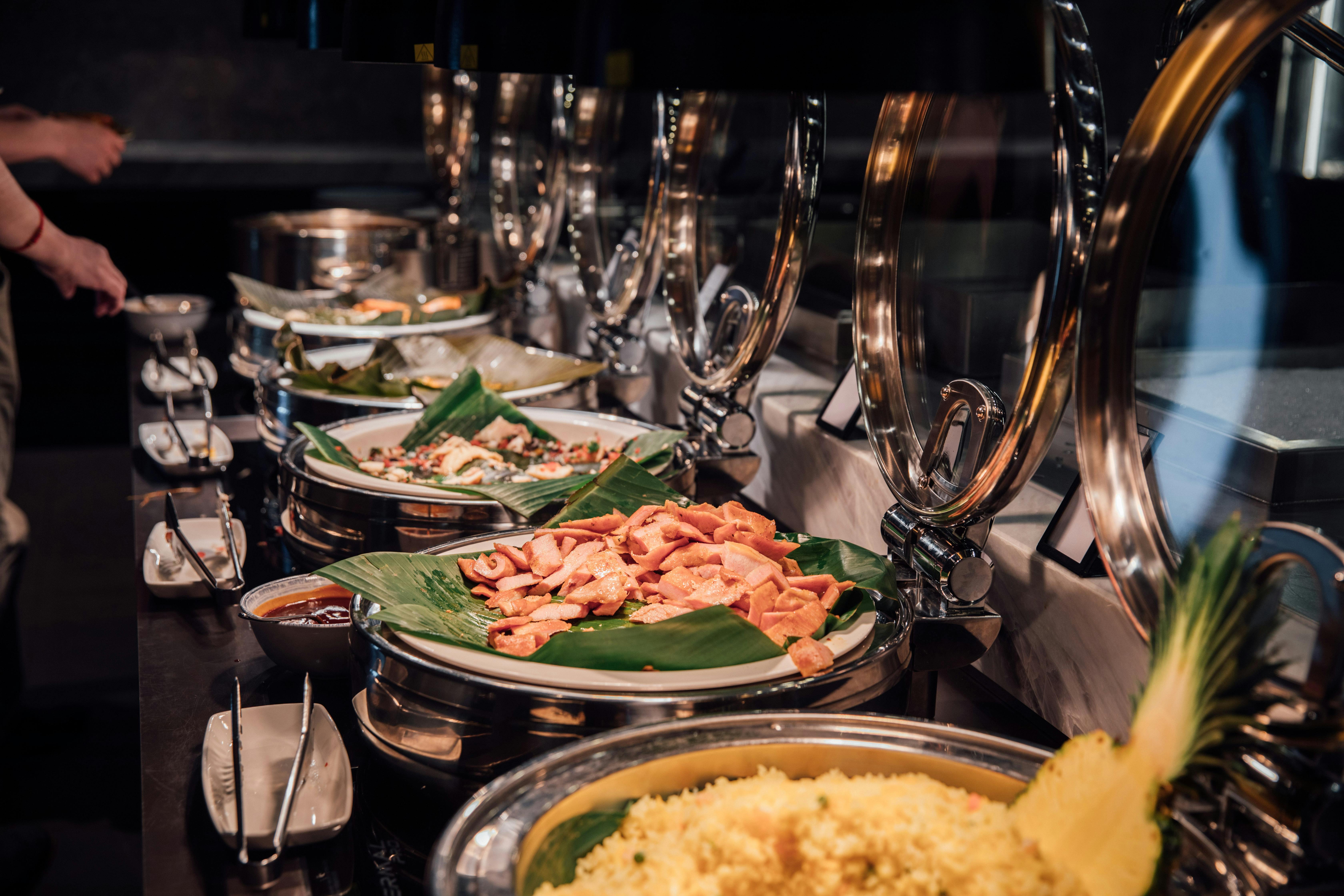 A long buffet line featuring stainless steel chafing dishes filled with food served on banana leaves, illustrating multi-site contract catering food service.