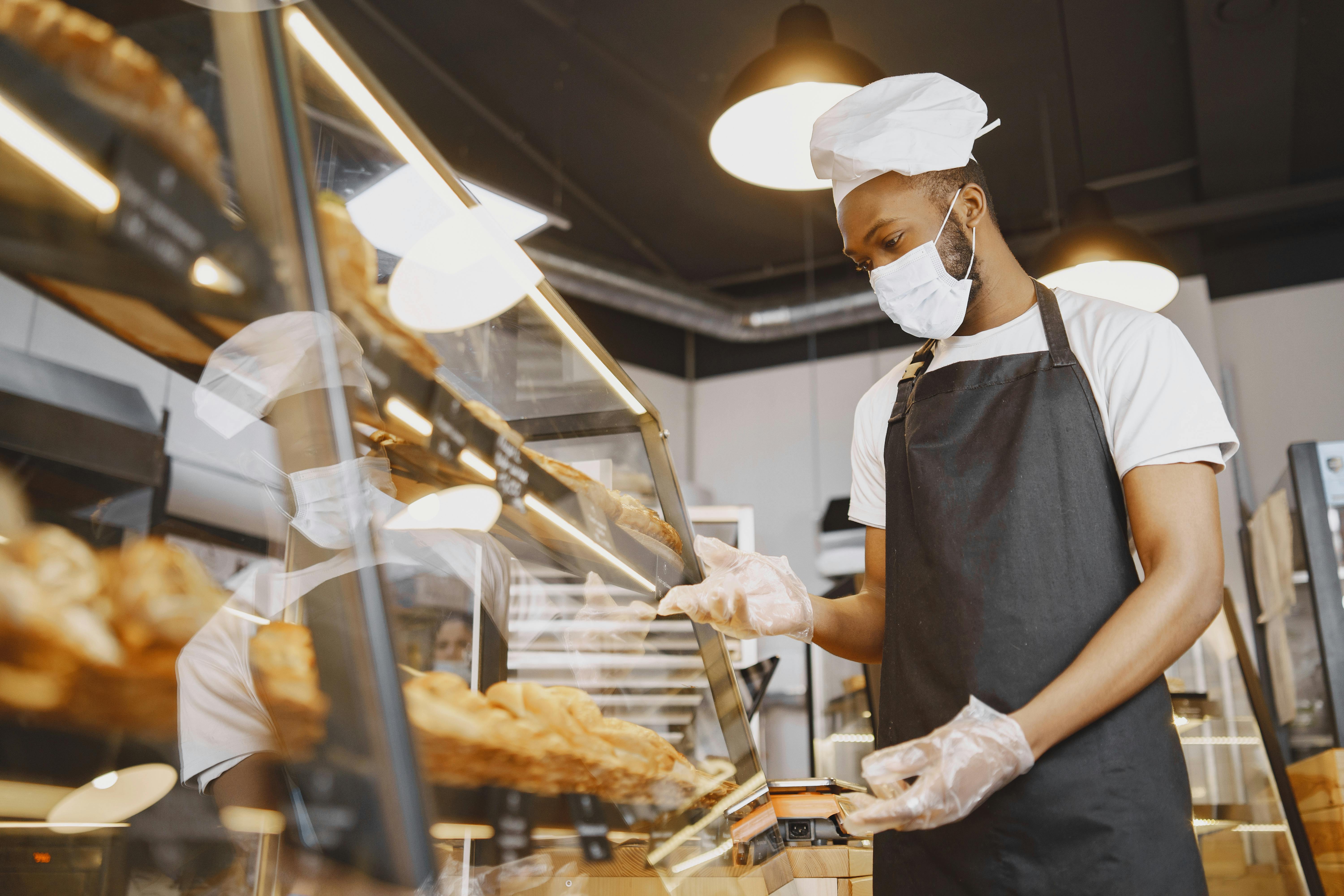 A bakery or food service worker wearing a white chef's hat, face mask, and plastic gloves inspecting a display case to maintain strict hygiene standards.