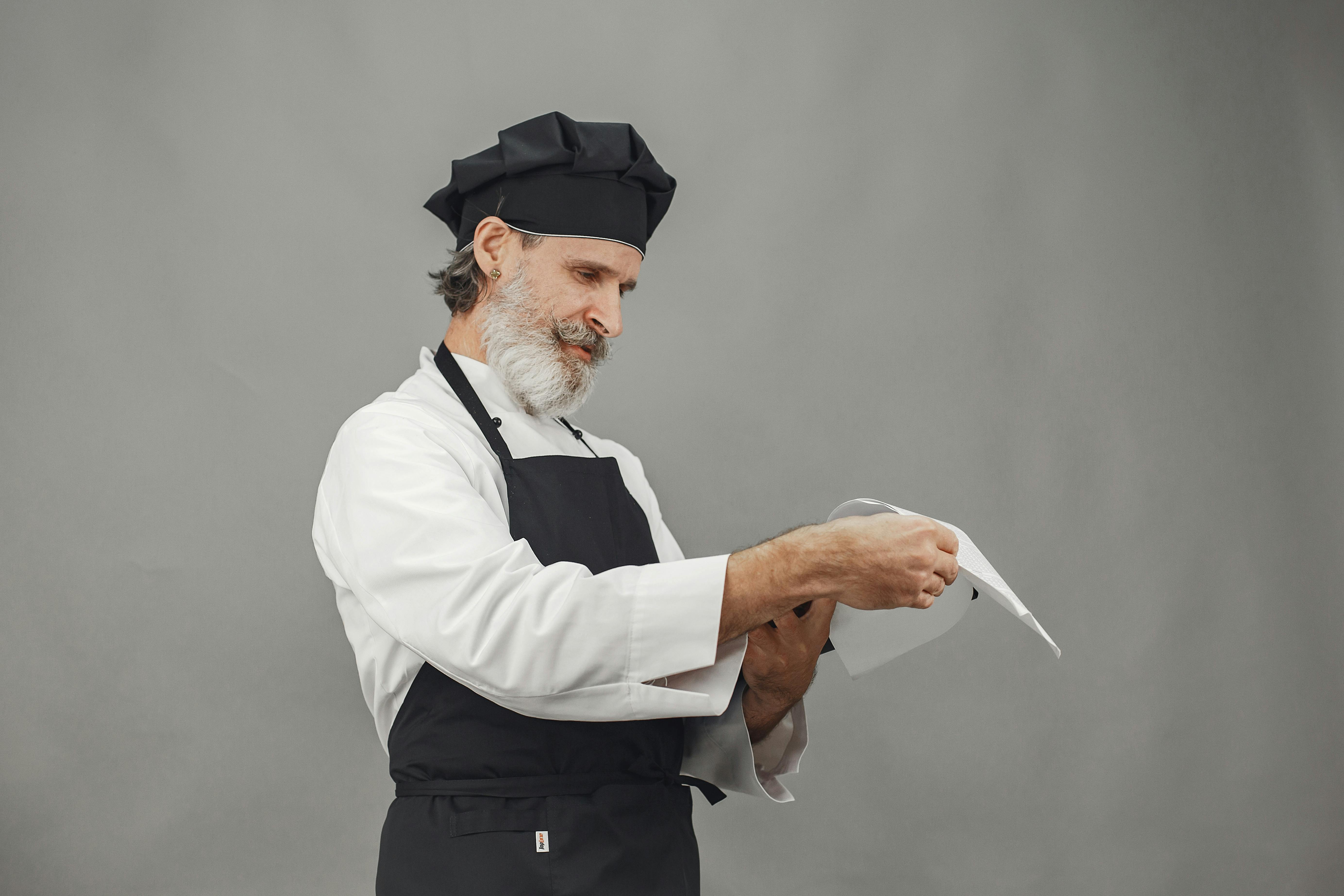 A professional chef wearing a white chef's hat, glasses, and blue hygienic gloves carefully plating food at a catering event buffet while guests look on.