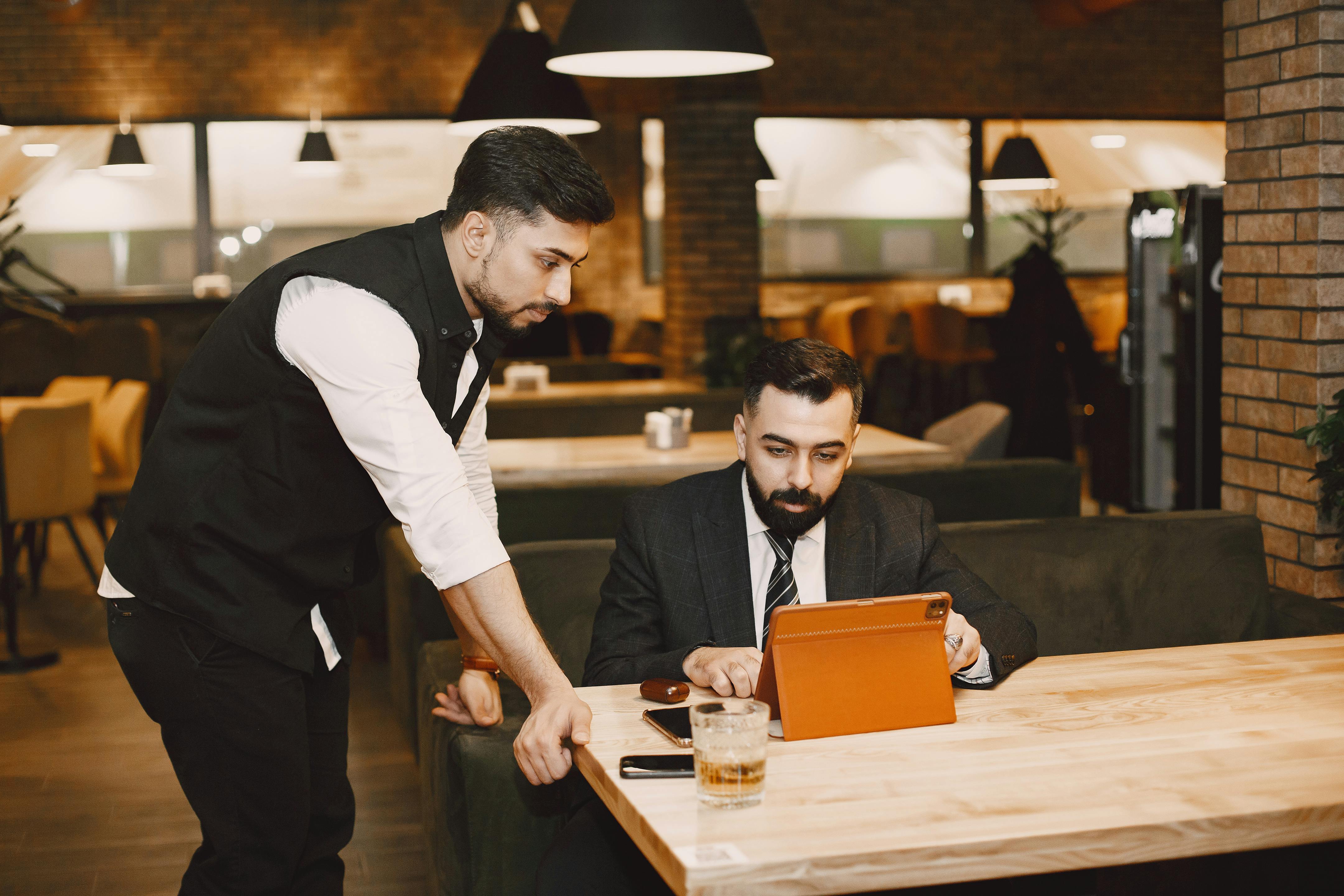 A restaurant or hotel manager in a business suit sitting at a wooden table and reviewing digital compliance data on a tablet while a floor supervisor or waiter in a vest stands beside him to discuss operations.