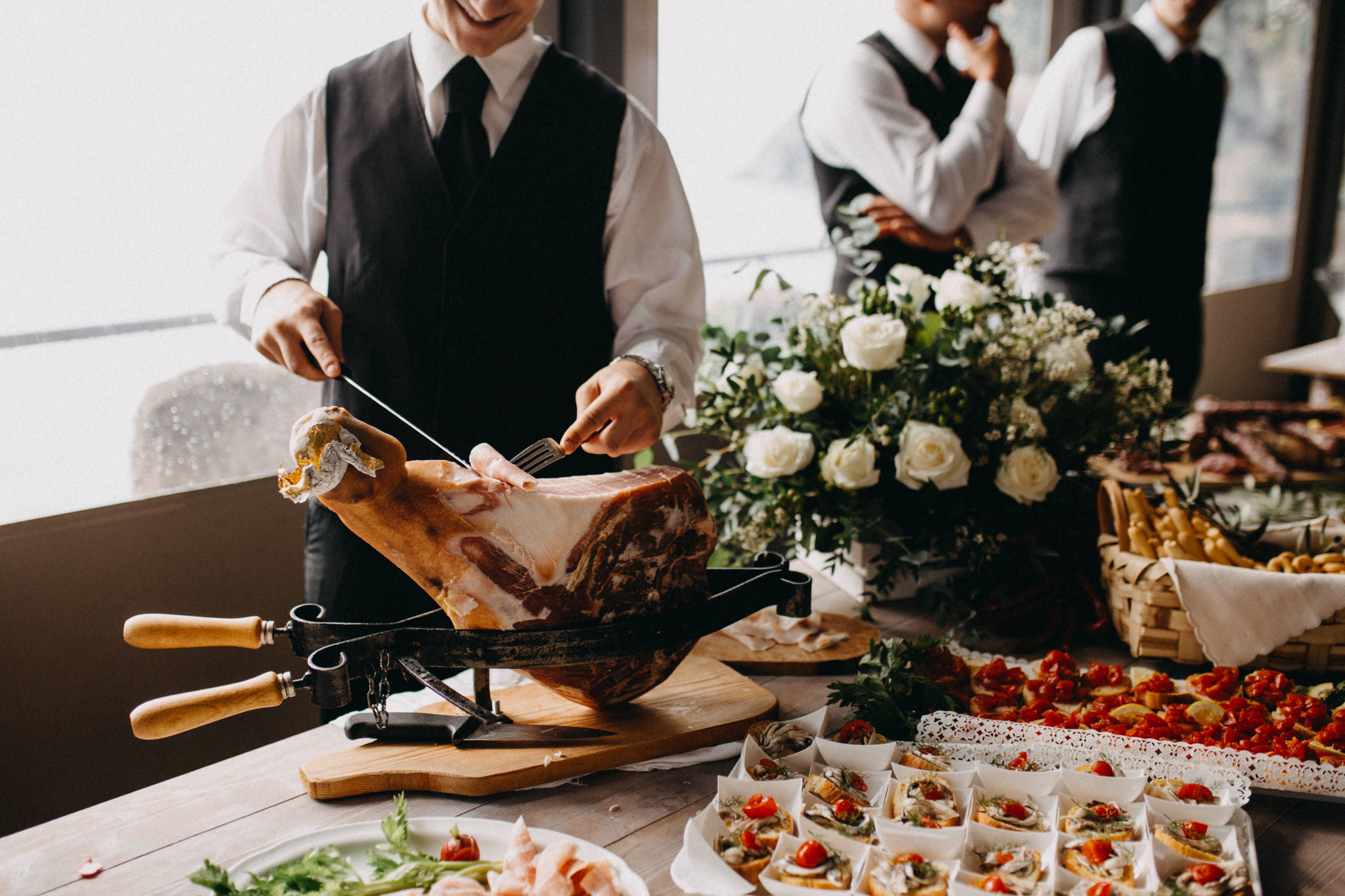 A professional waiter slicing a large cured ham leg on a wooden board at a beautifully catered buffet table filled with gourmet appetizers and fresh white roses.
