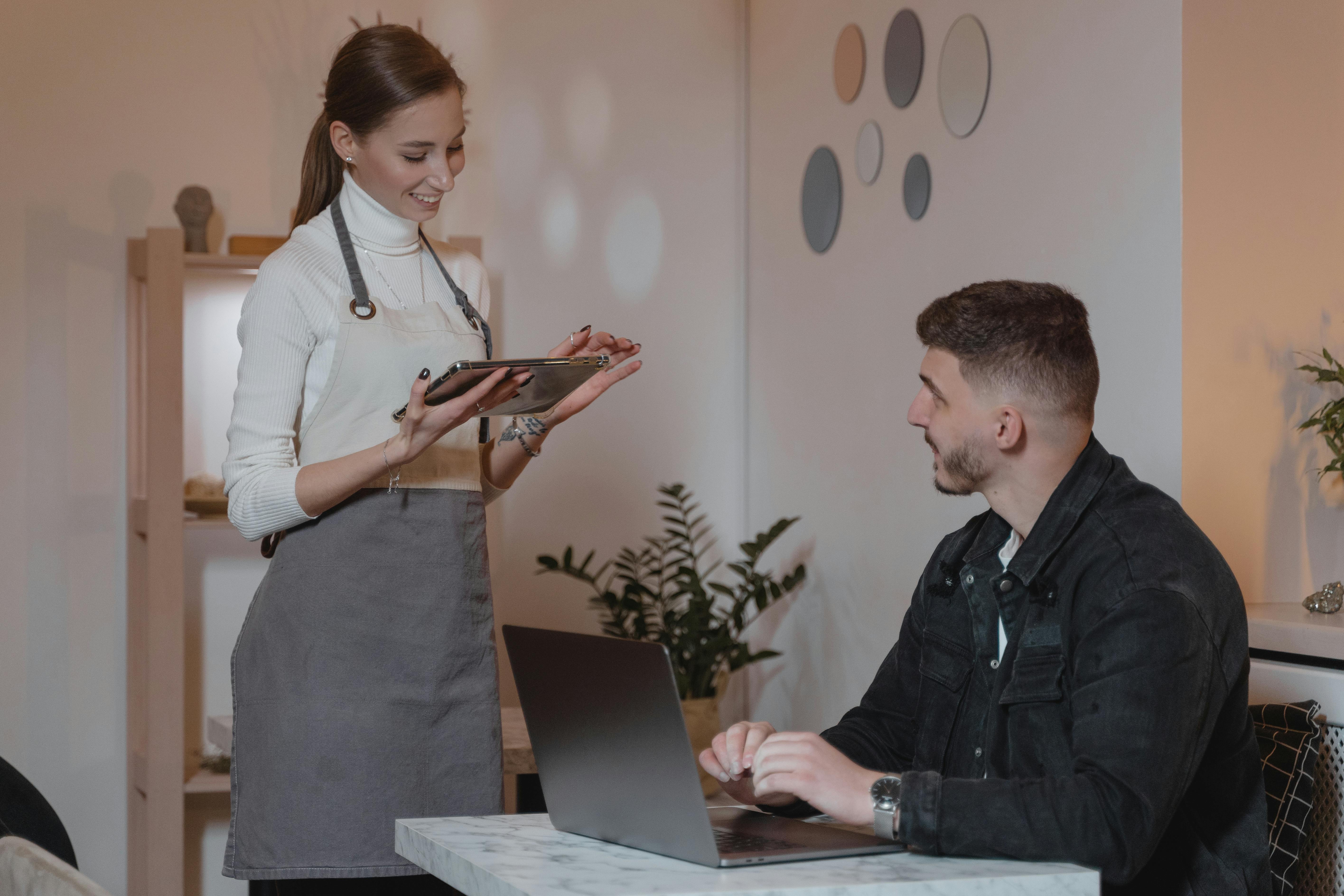 A friendly female server wearing an apron holds a digital tablet while taking an order from a male customer sitting at a table with his laptop in a modern café setting.