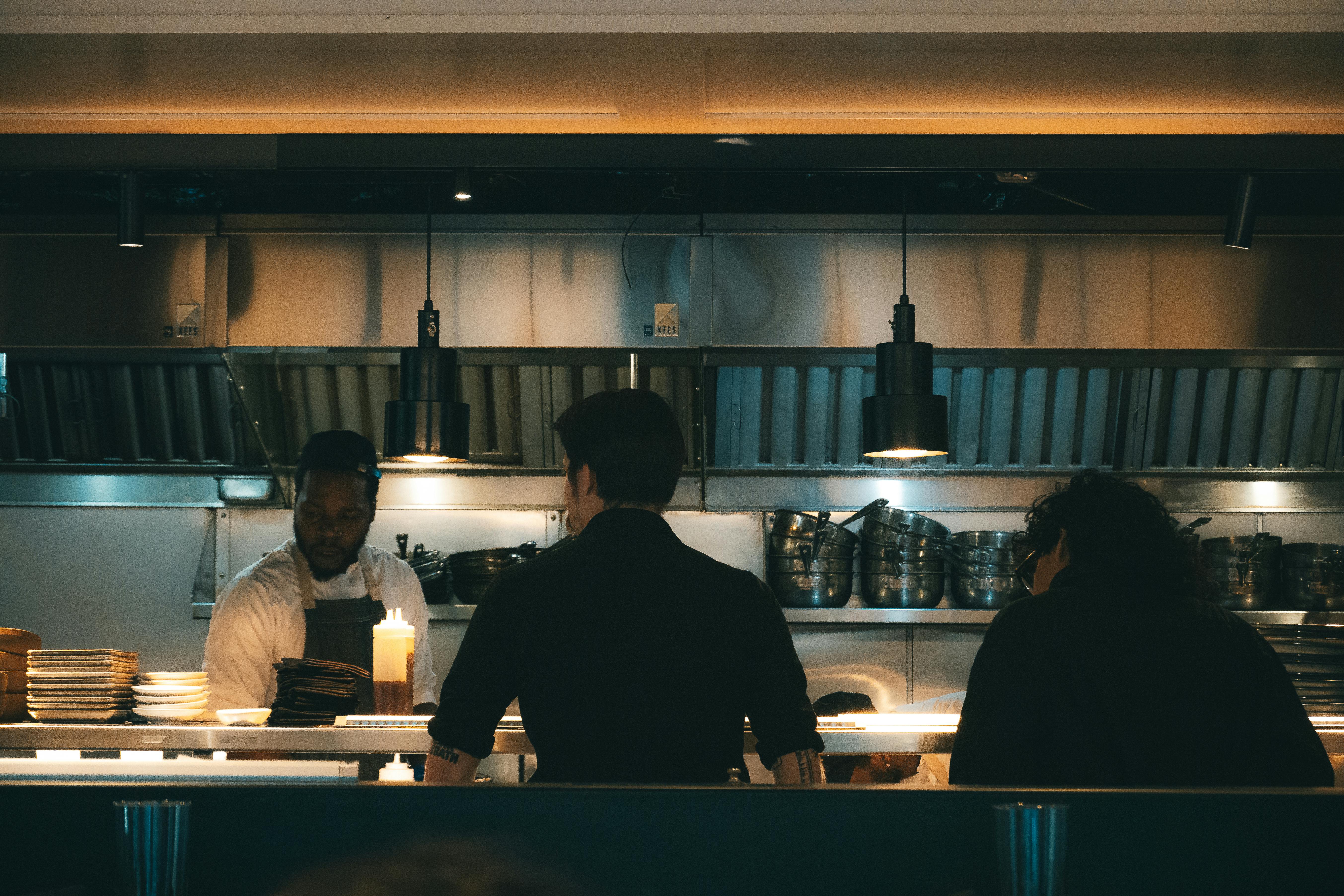 A dim, late-night professional commercial kitchen scene showing three kitchen staff members working behind the pass counter under warm hanging lights.