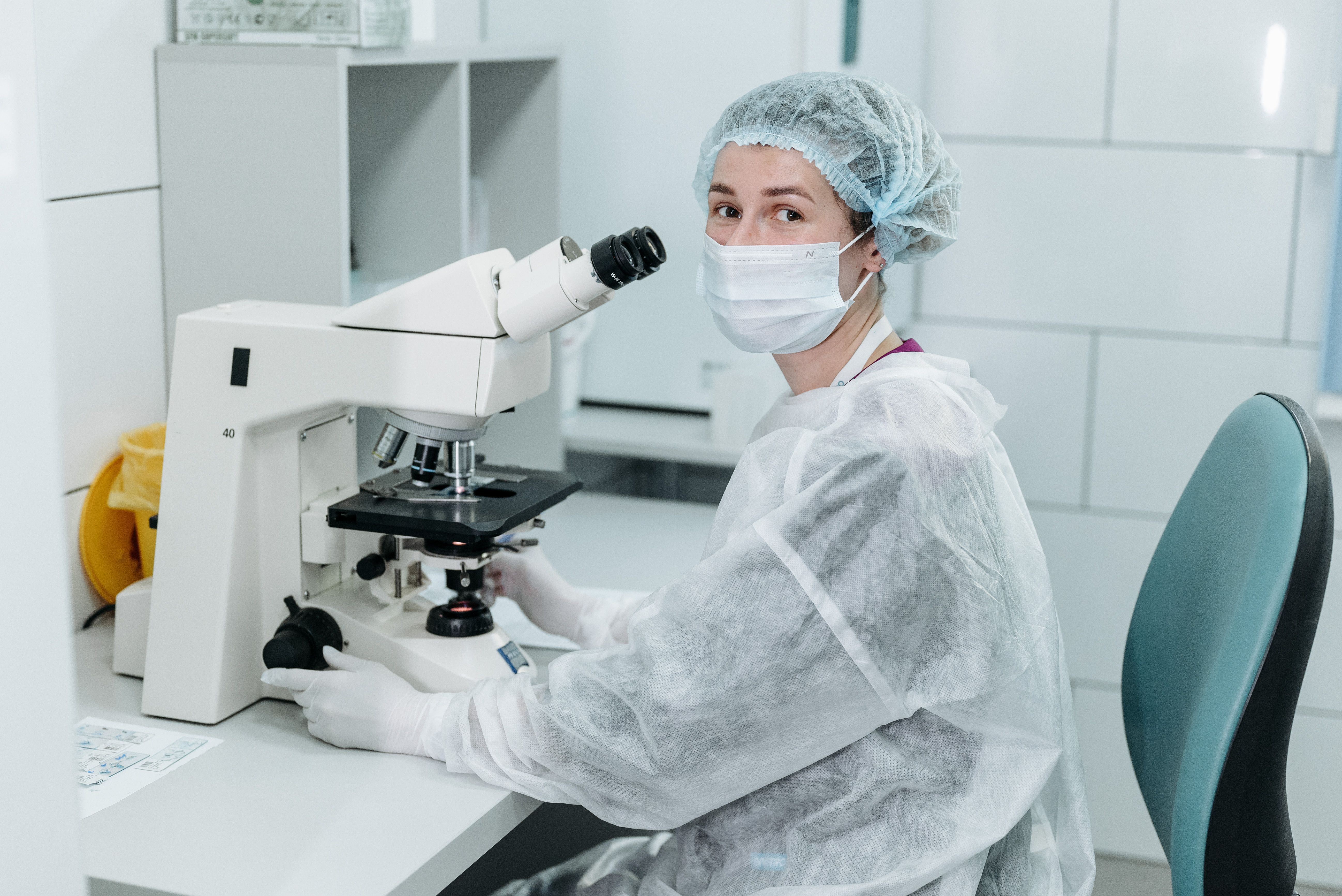 Laboratory technician in protective gown and mask examining a sample under a microscope