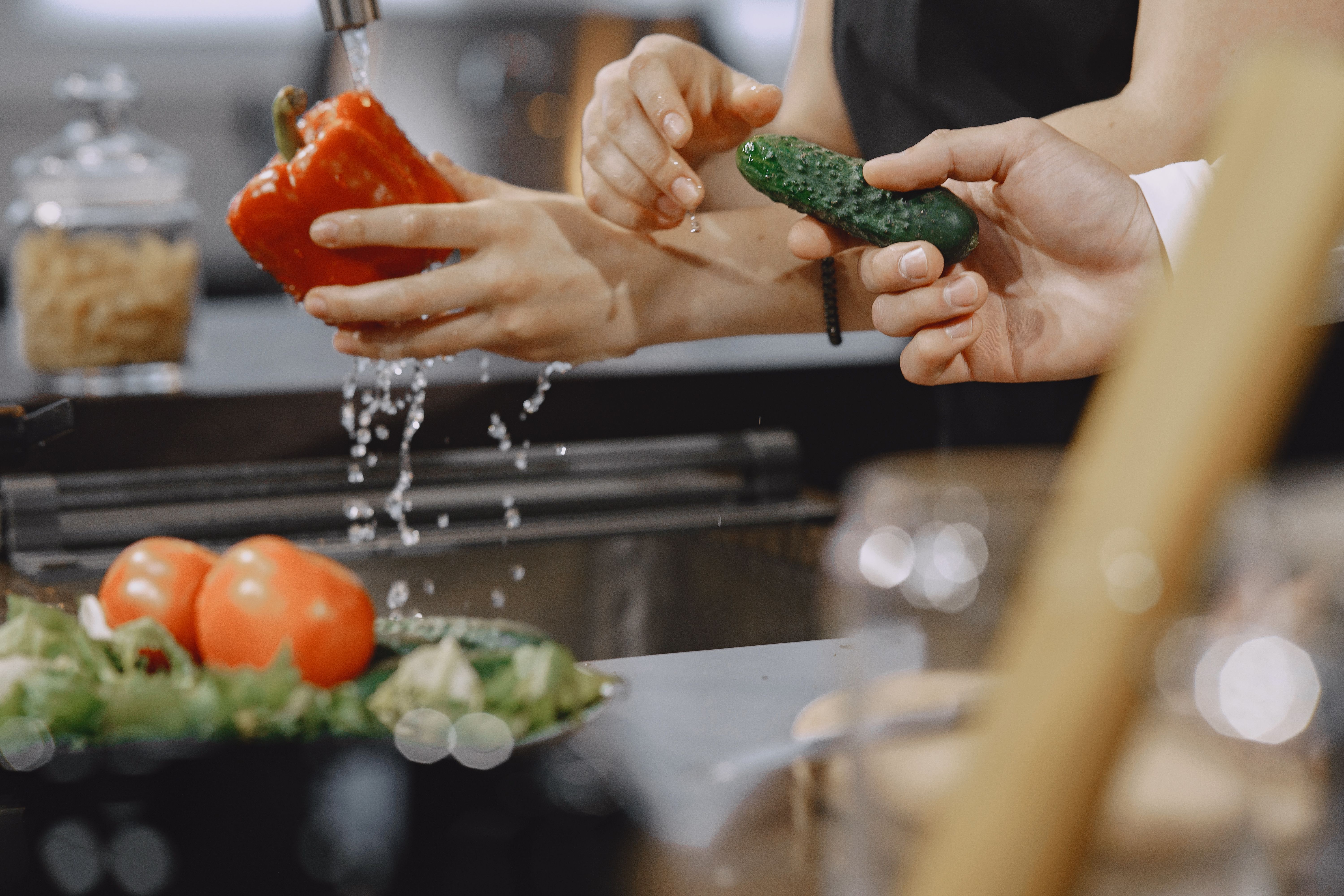 Hands rinsing a red pepper and cucumber under running water at a kitchen sink