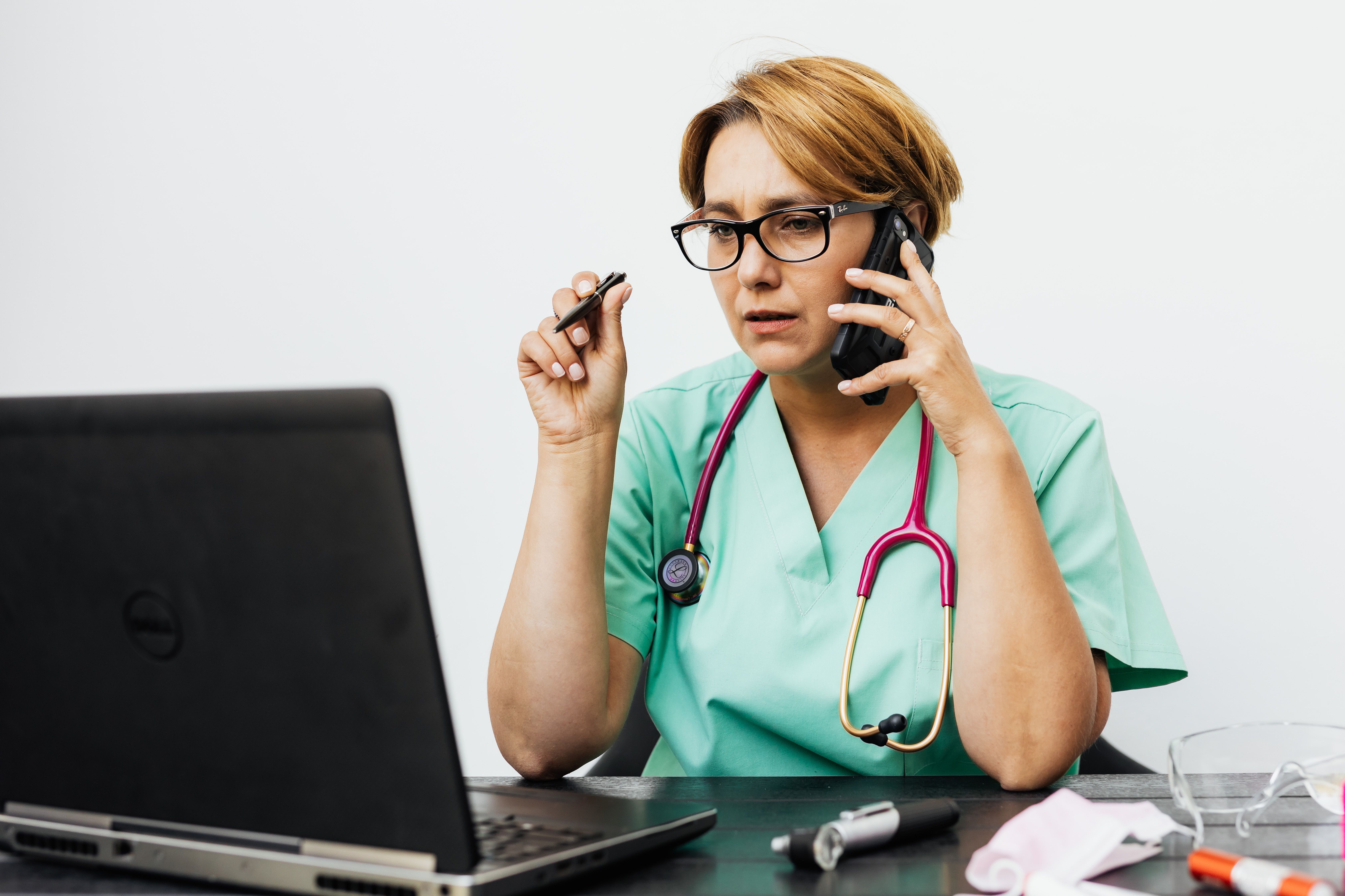 Nurse in scrubs with a stethoscope speaking on the phone while reviewing information on a laptop