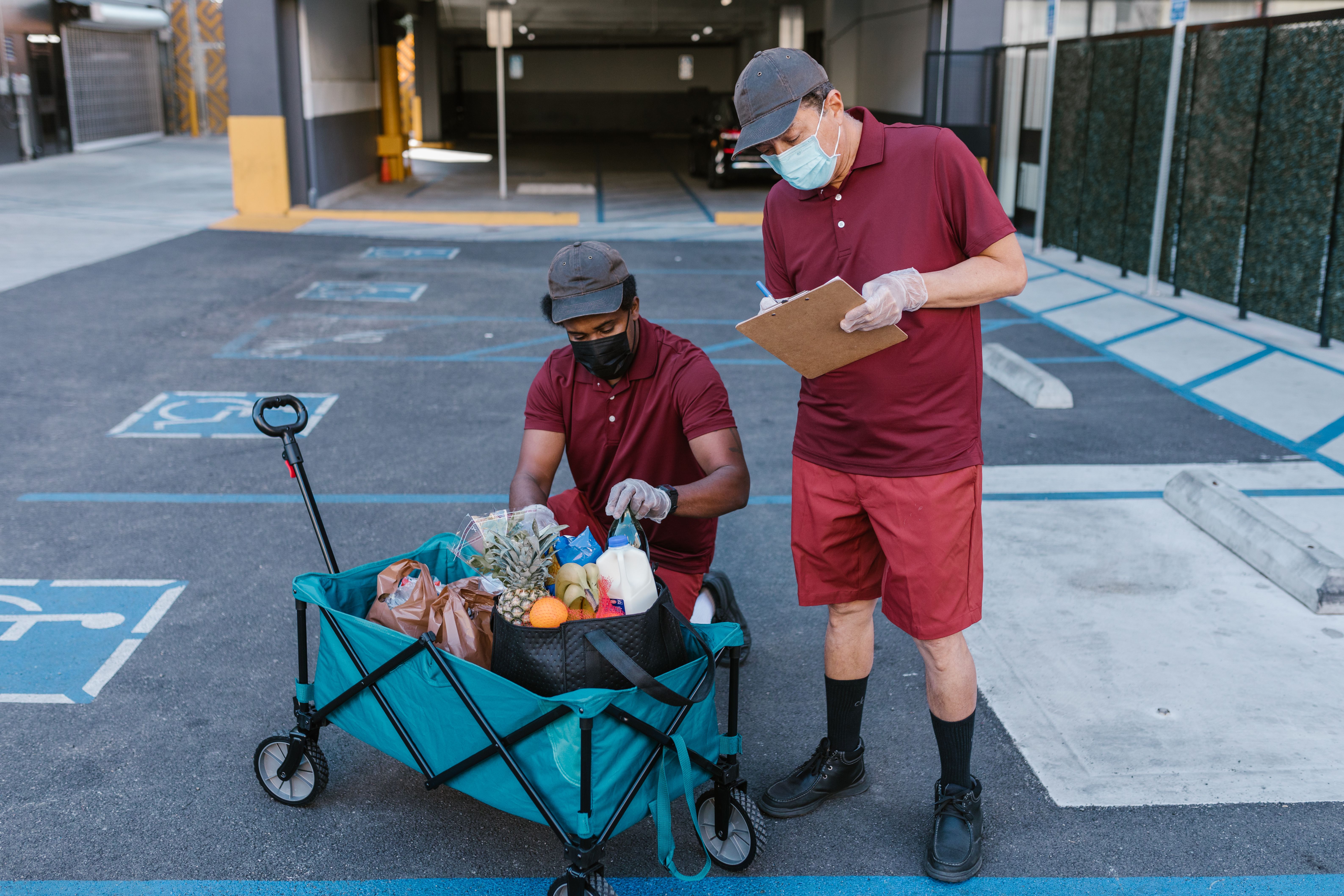 Delivery personnel in protective gear checking a grocery inventory list in a parking area.