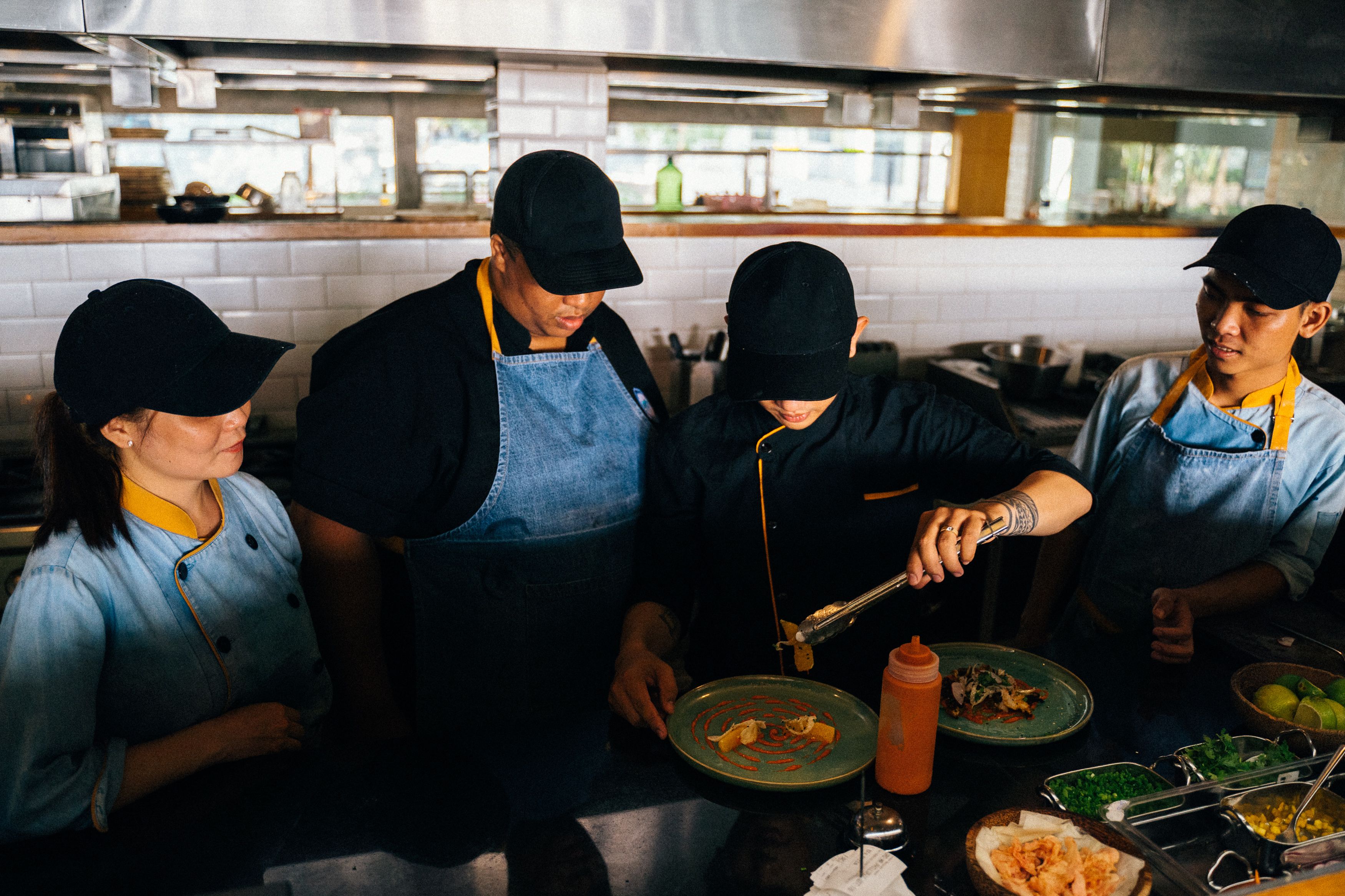 A group of four kitchen staff members in matching uniforms and caps carefully plating dishes on a commercial kitchen counter.