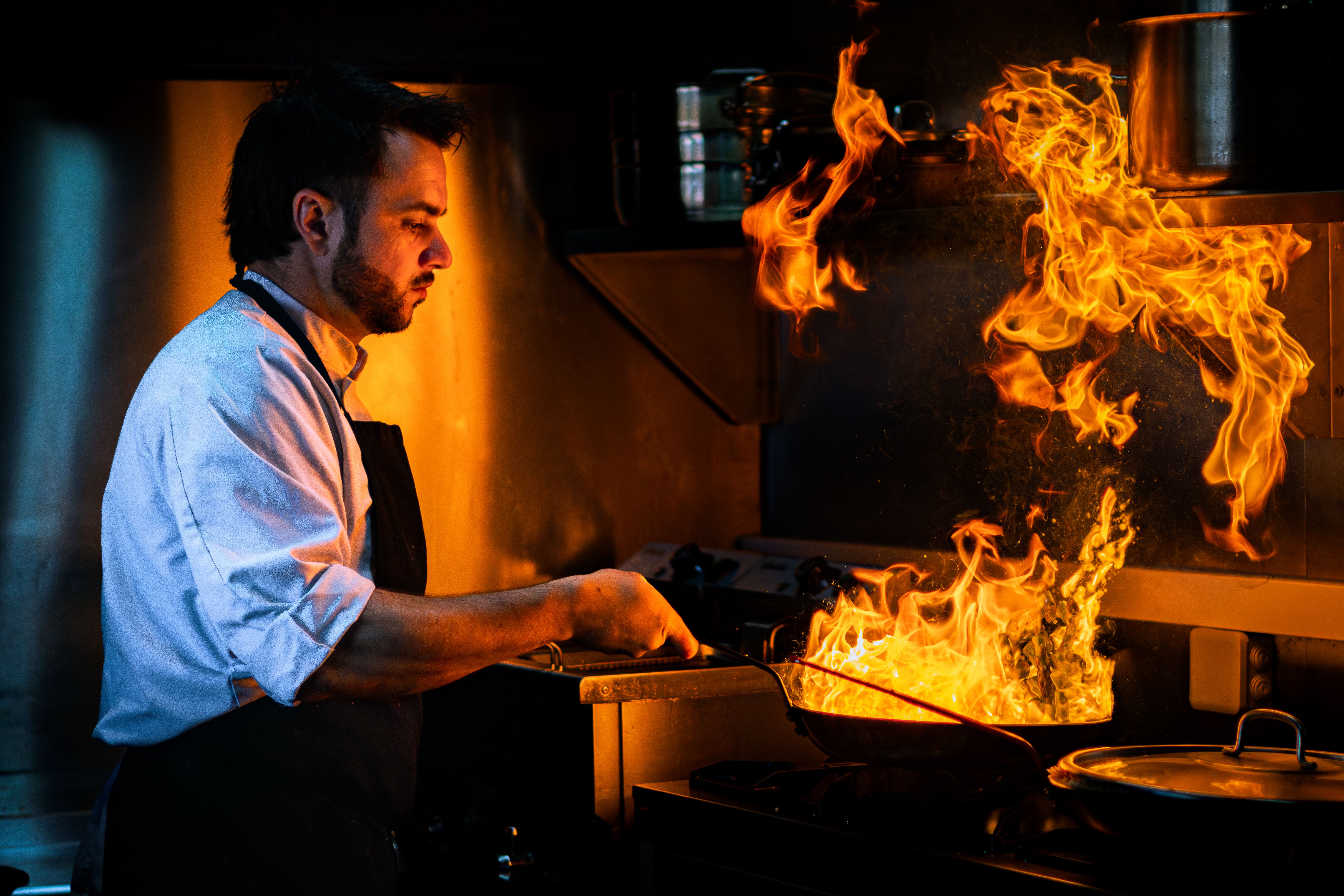A male chef in an apron stirring food in a pan with large dramatic flames flaring up on a commercial stove.