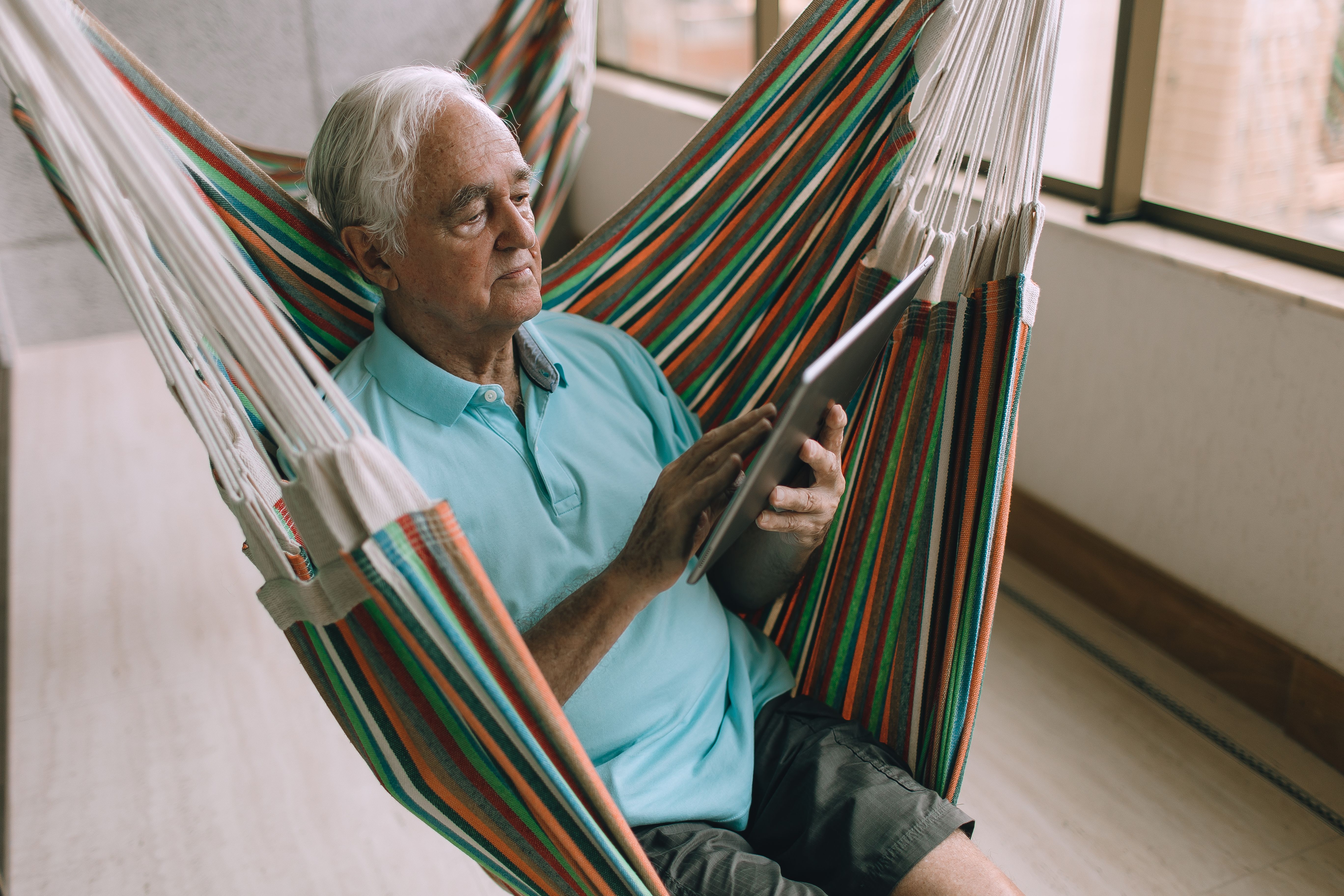 An elderly man sitting comfortably in a colorful striped hammock while interacting with a digital tablet.