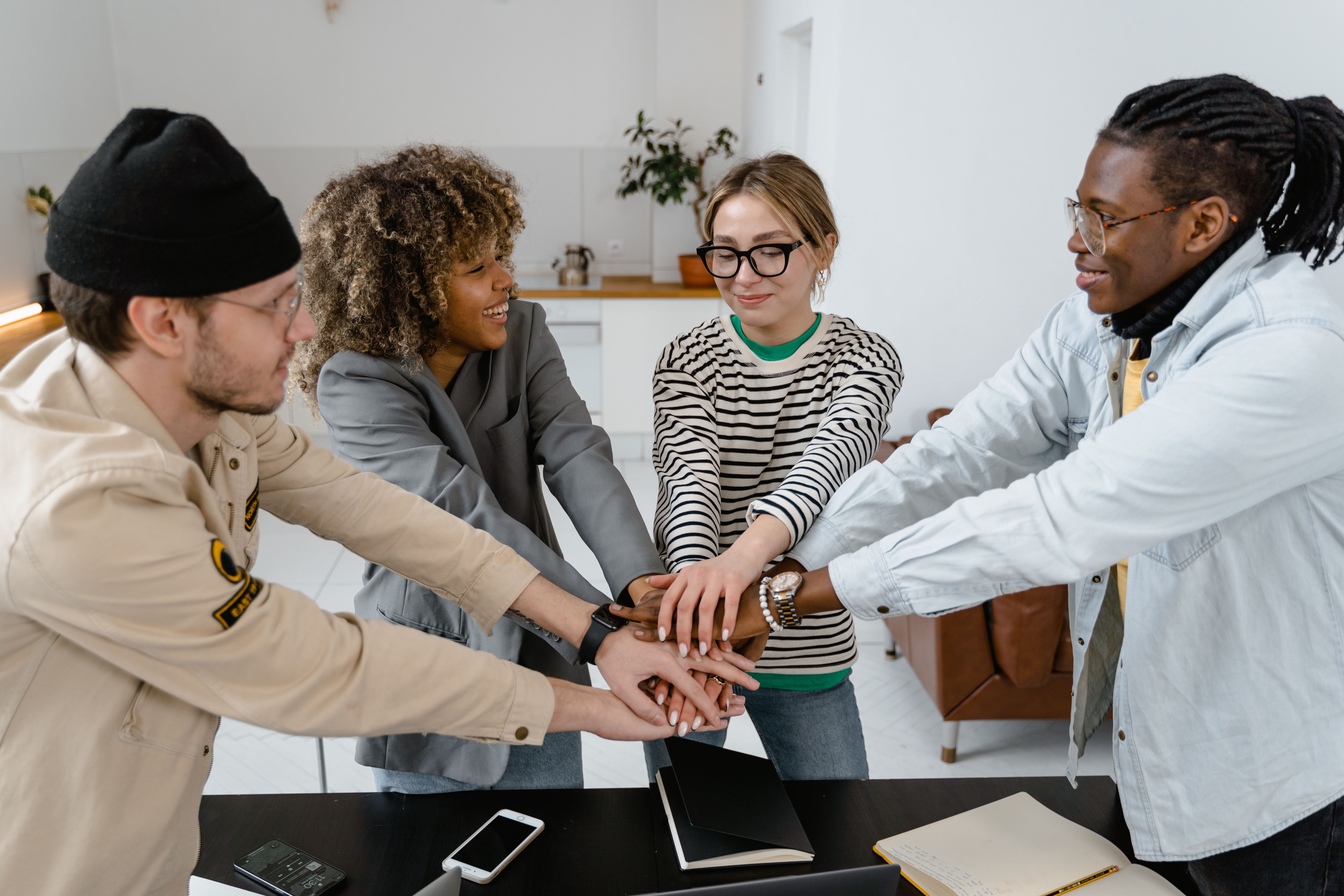 A diverse team of four colleagues standing around a desk with hands stacked together in a high-five gesture of teamwork in a modern office space.