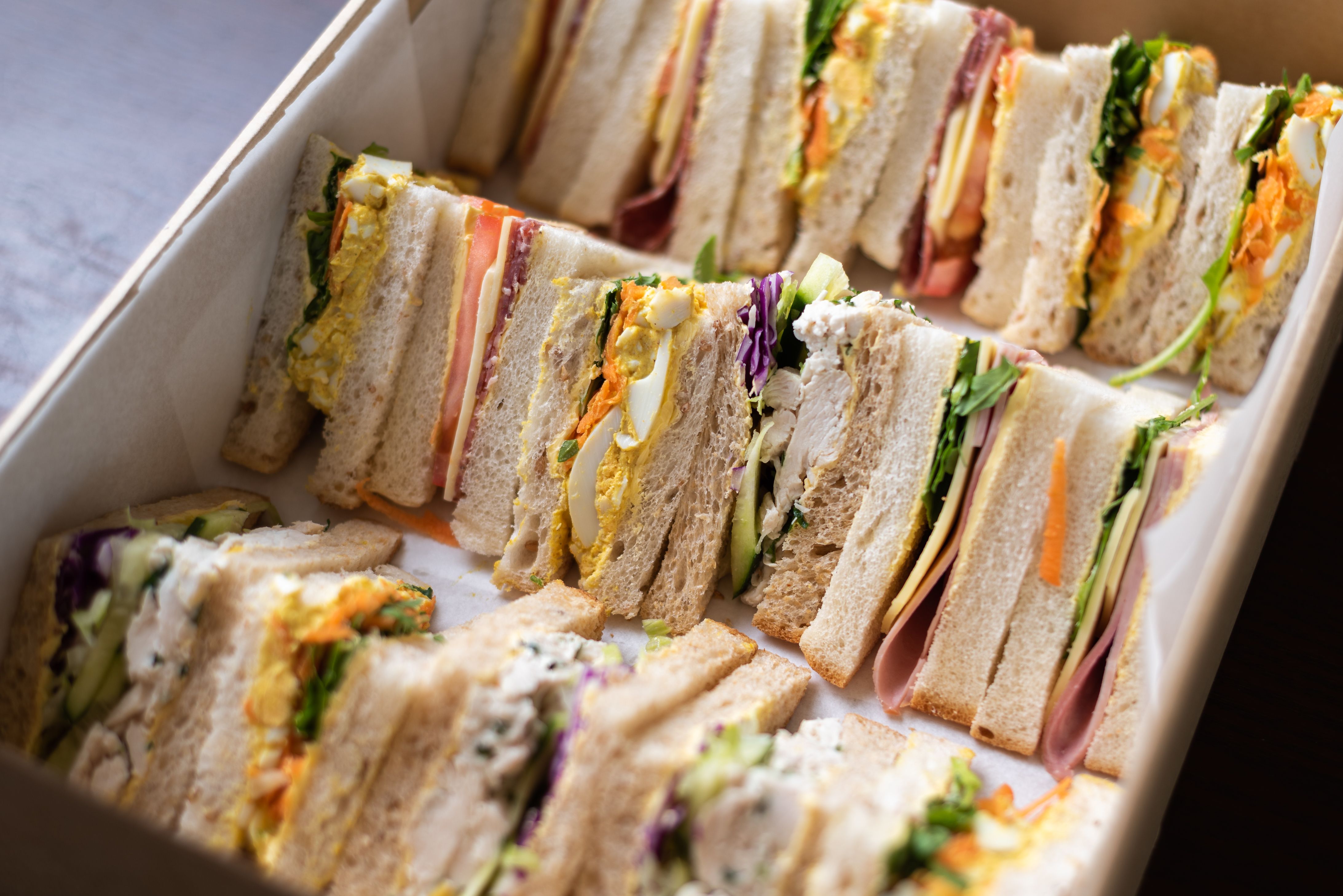 An assortment of fresh, neatly cut triangular sandwiches with various fillings packaged inside a cardboard catering box.