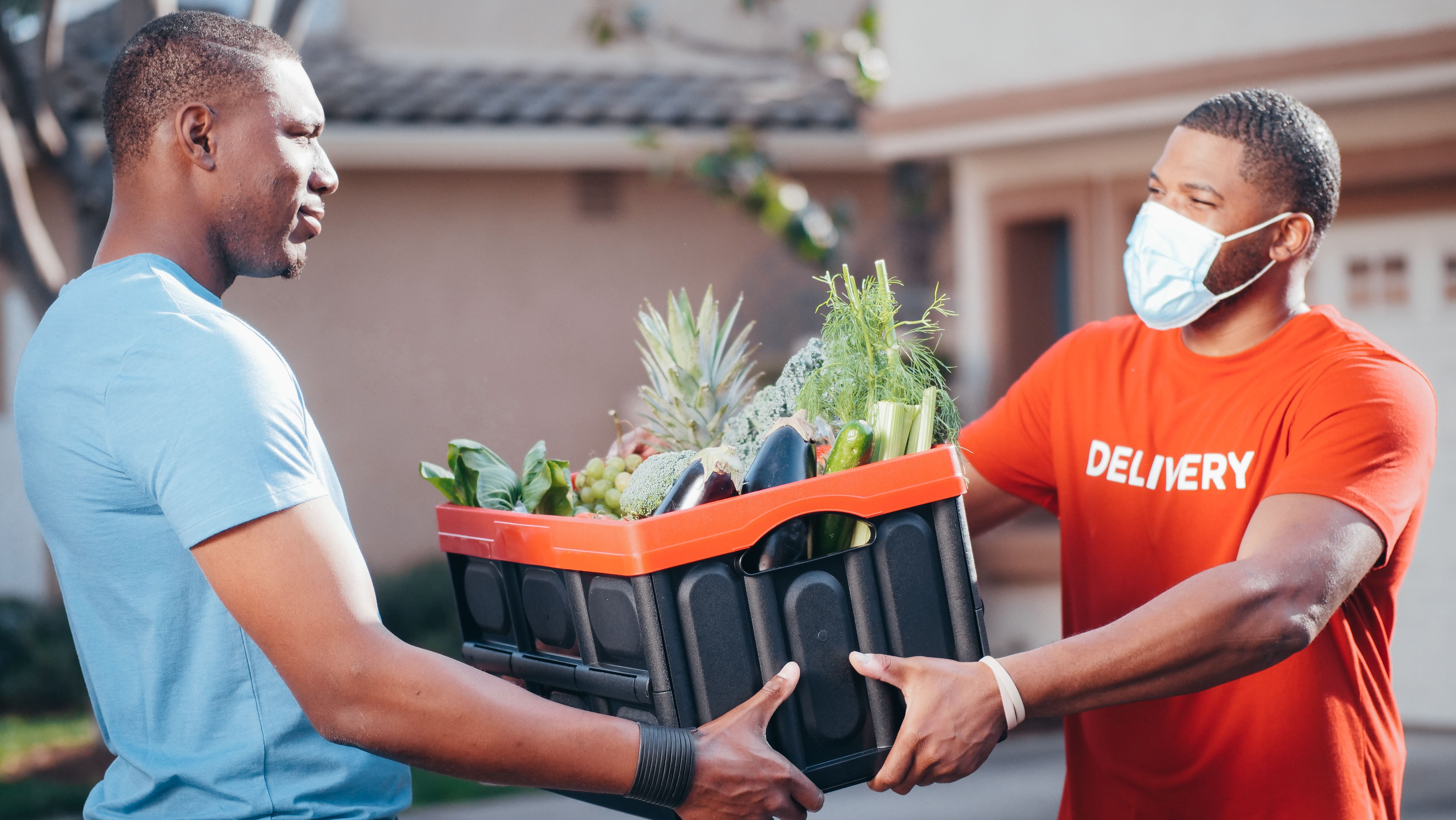 A delivery worker in a red shirt and face mask handing a crate full of fresh groceries to a customer outside a house.