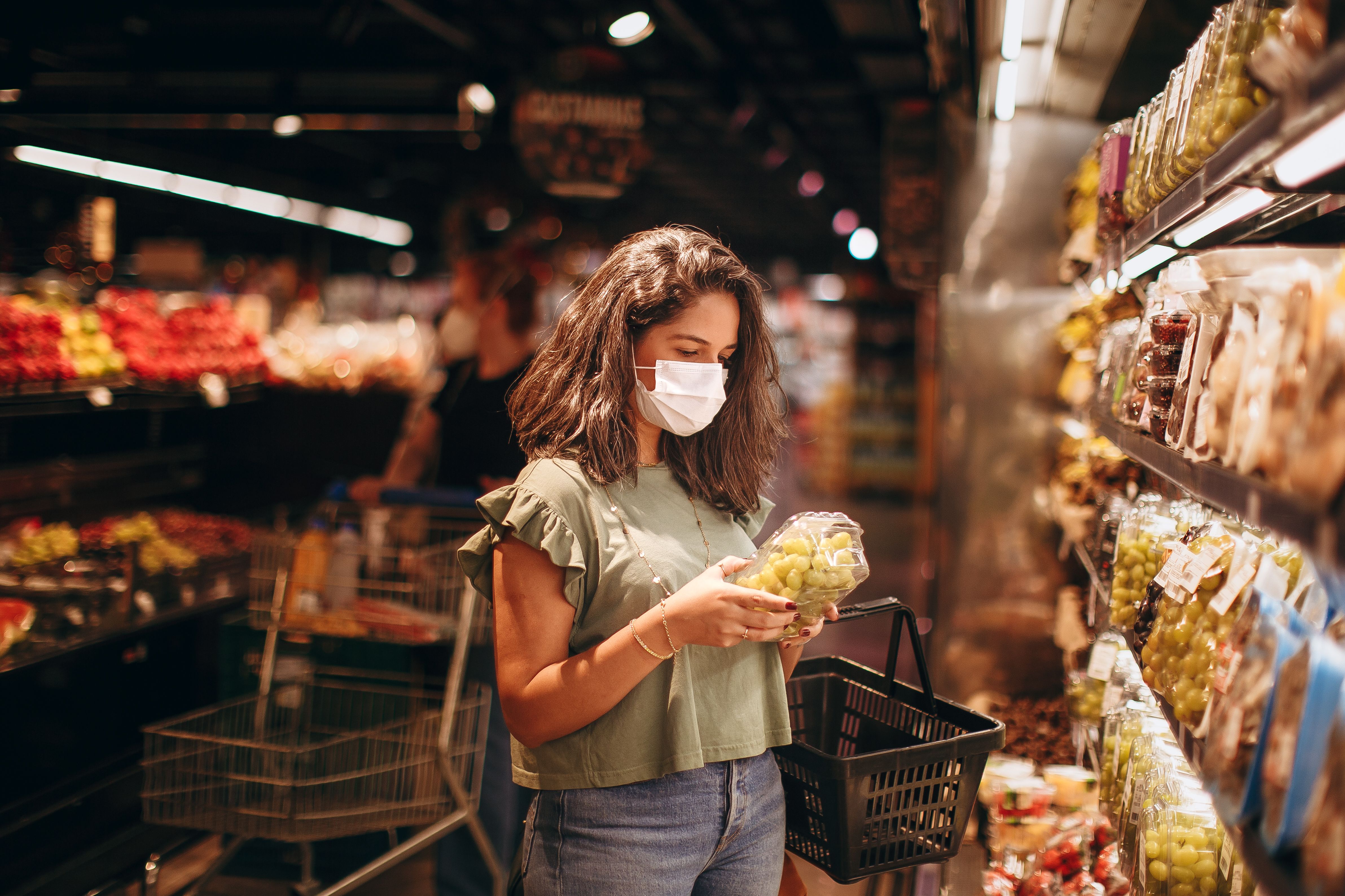 A woman wearing a protective face mask examining a plastic container of green grapes in a brightly lit grocery store aisle.