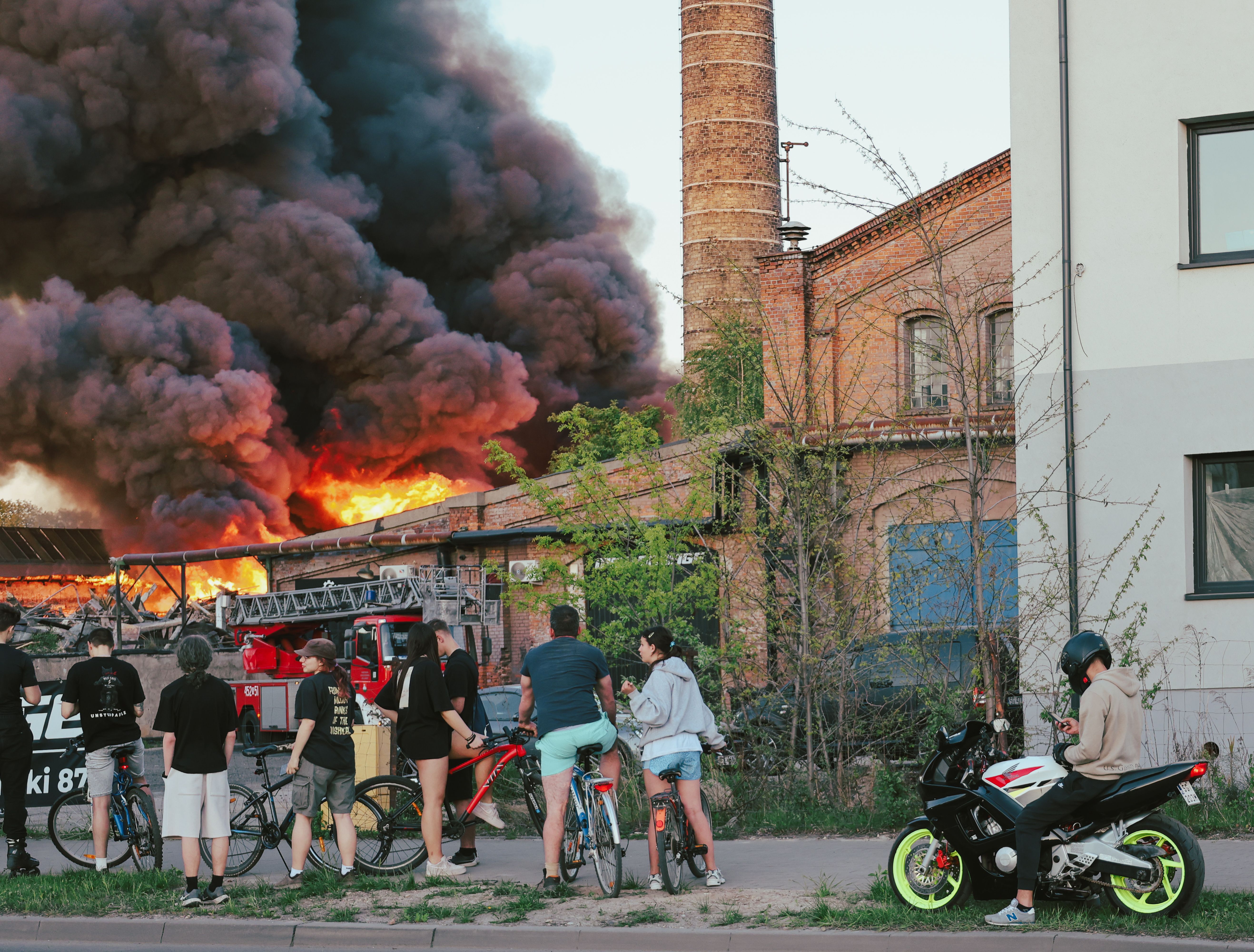 A group of bystanders and cyclists watching a massive building fire with thick dark smoke and a fire truck in the background.