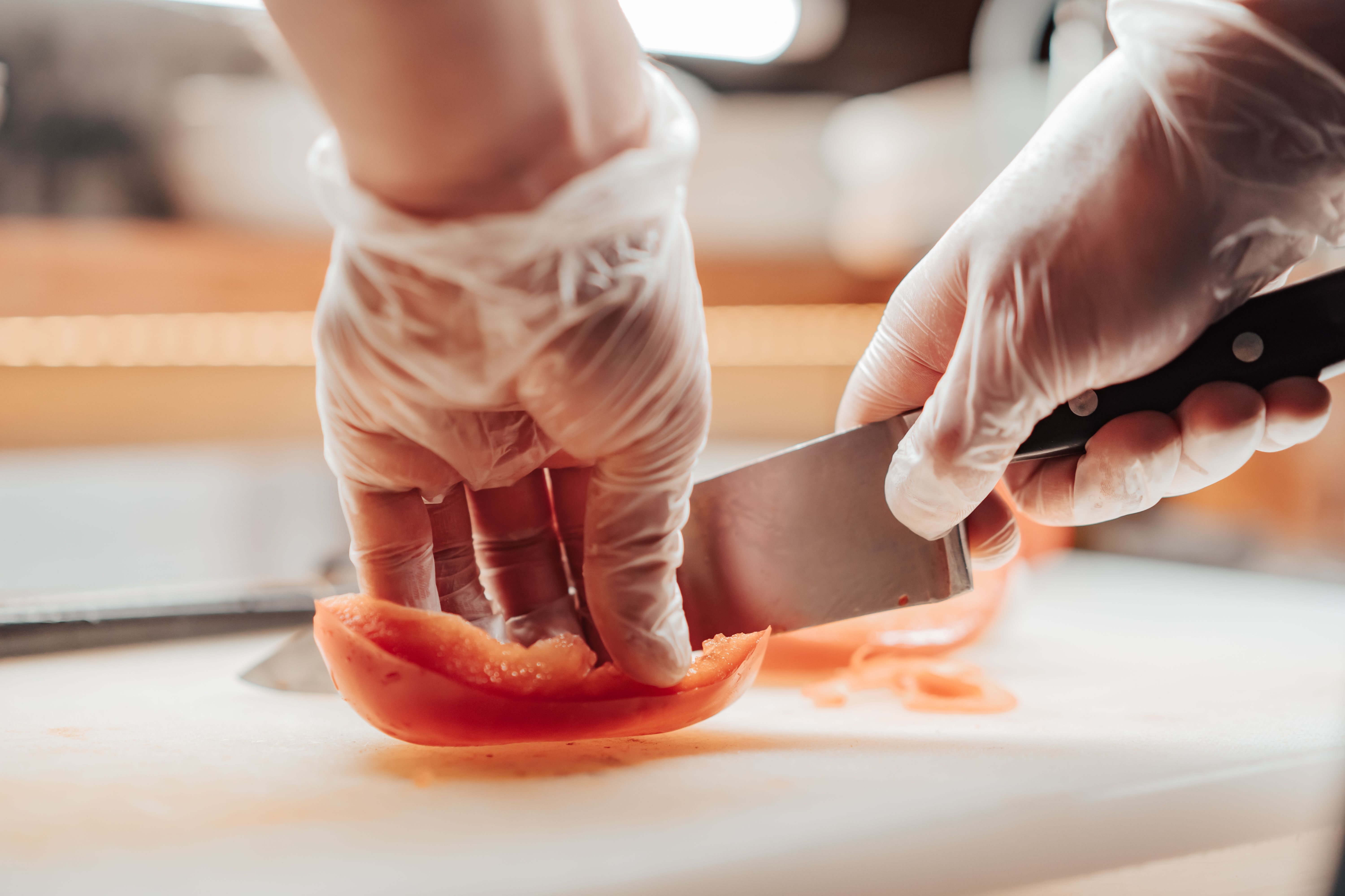 A close-up of hands wearing clear hygiene gloves slicing a red bell pepper with a sharp knife on a cutting board.