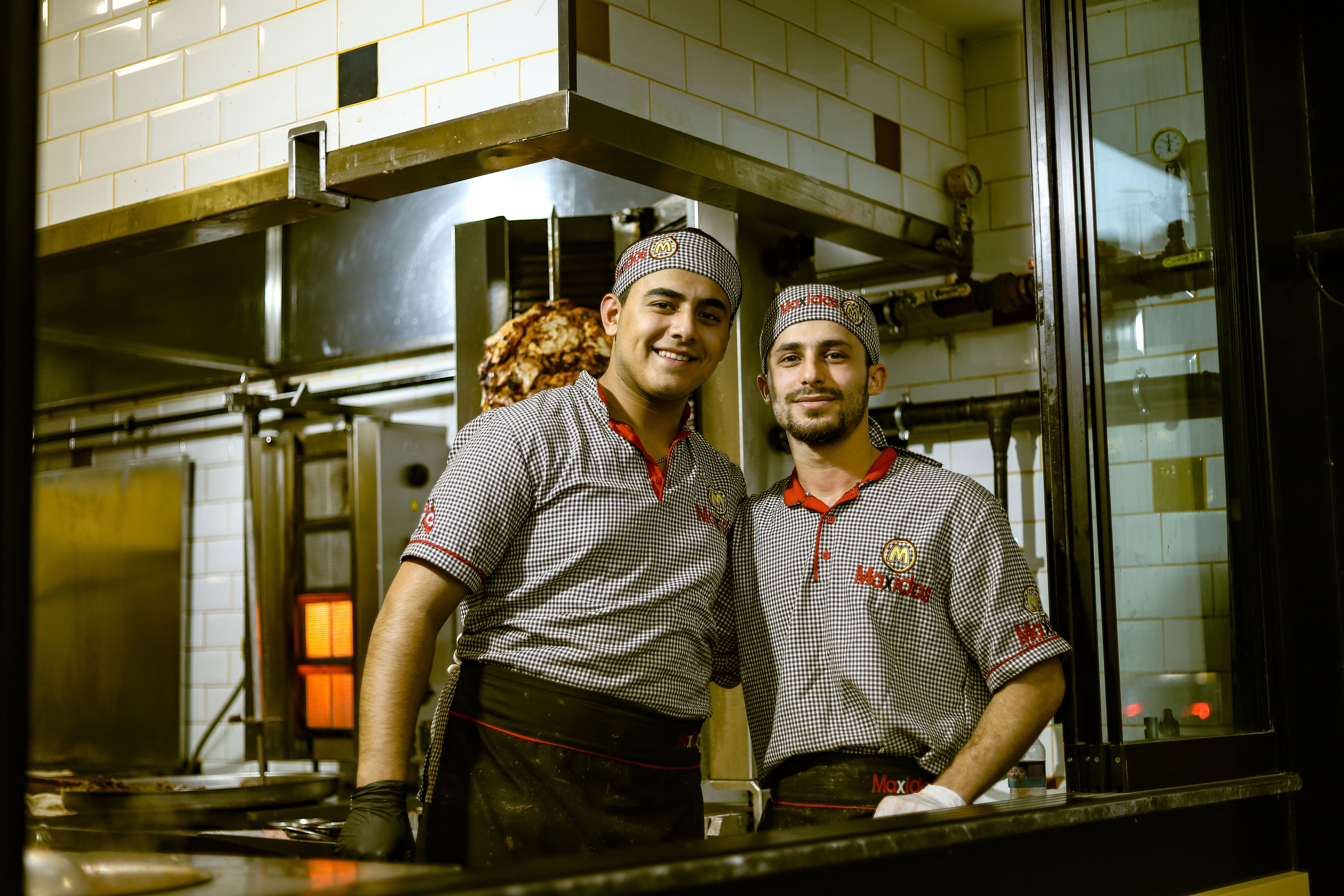 Two smiling male kitchen staff members wearing matching patterned shirts and hats standing together in a restaurant kitchen.