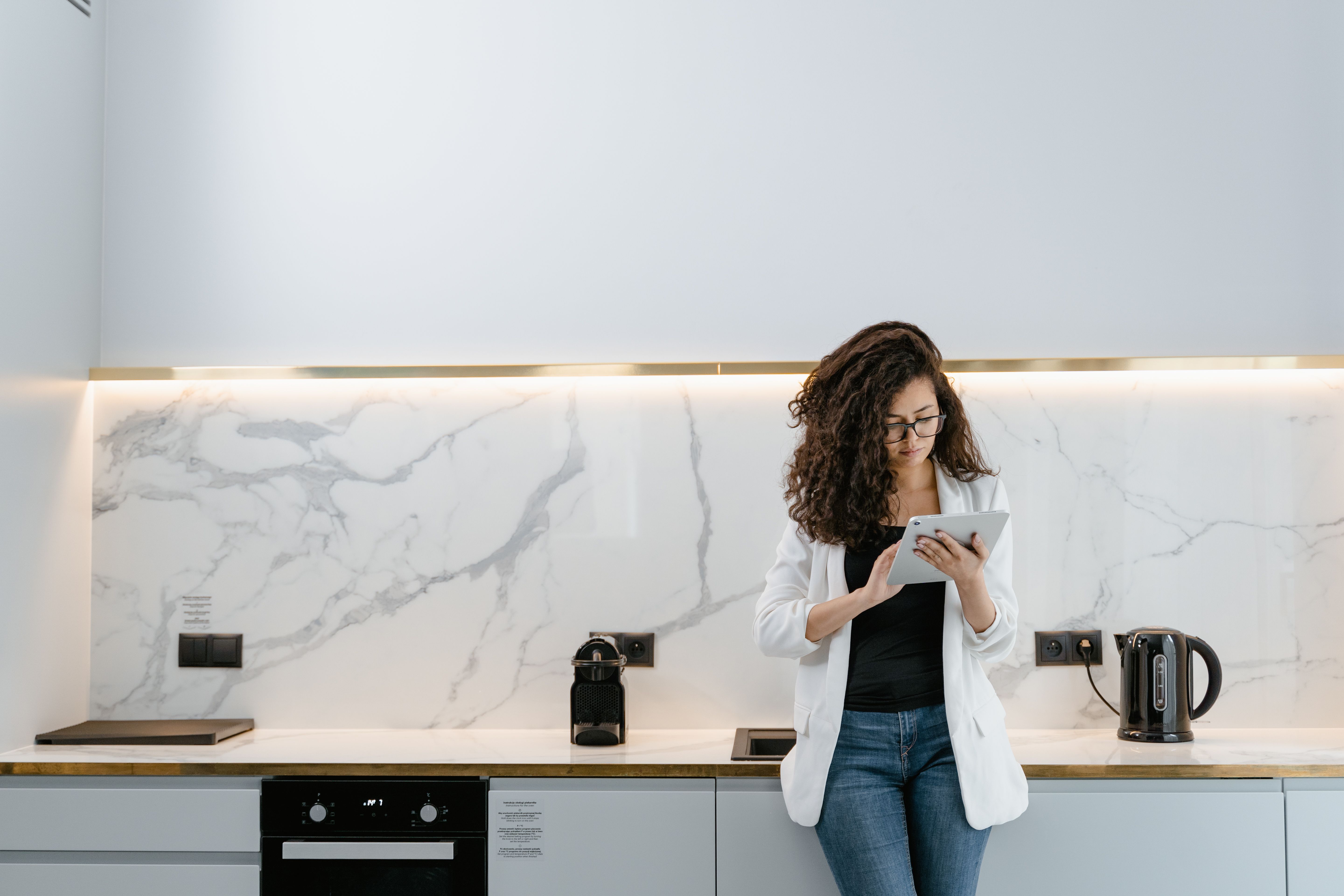 A woman in a white blazer and glasses using a digital tablet while standing in a modern kitchen with marble backsplash.