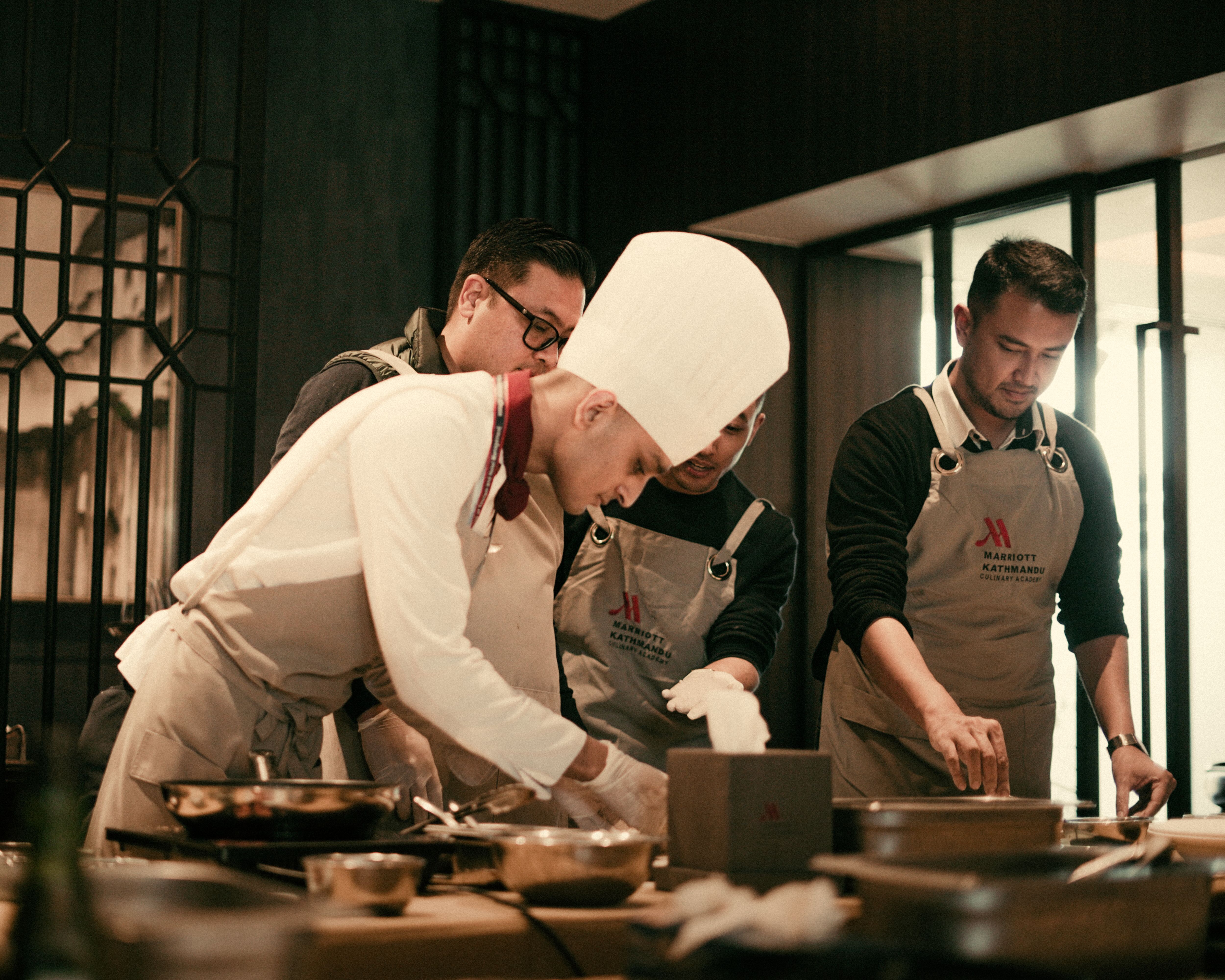 A group of chefs and kitchen staff wearing aprons preparing dishes together at a commercial kitchen counter.
