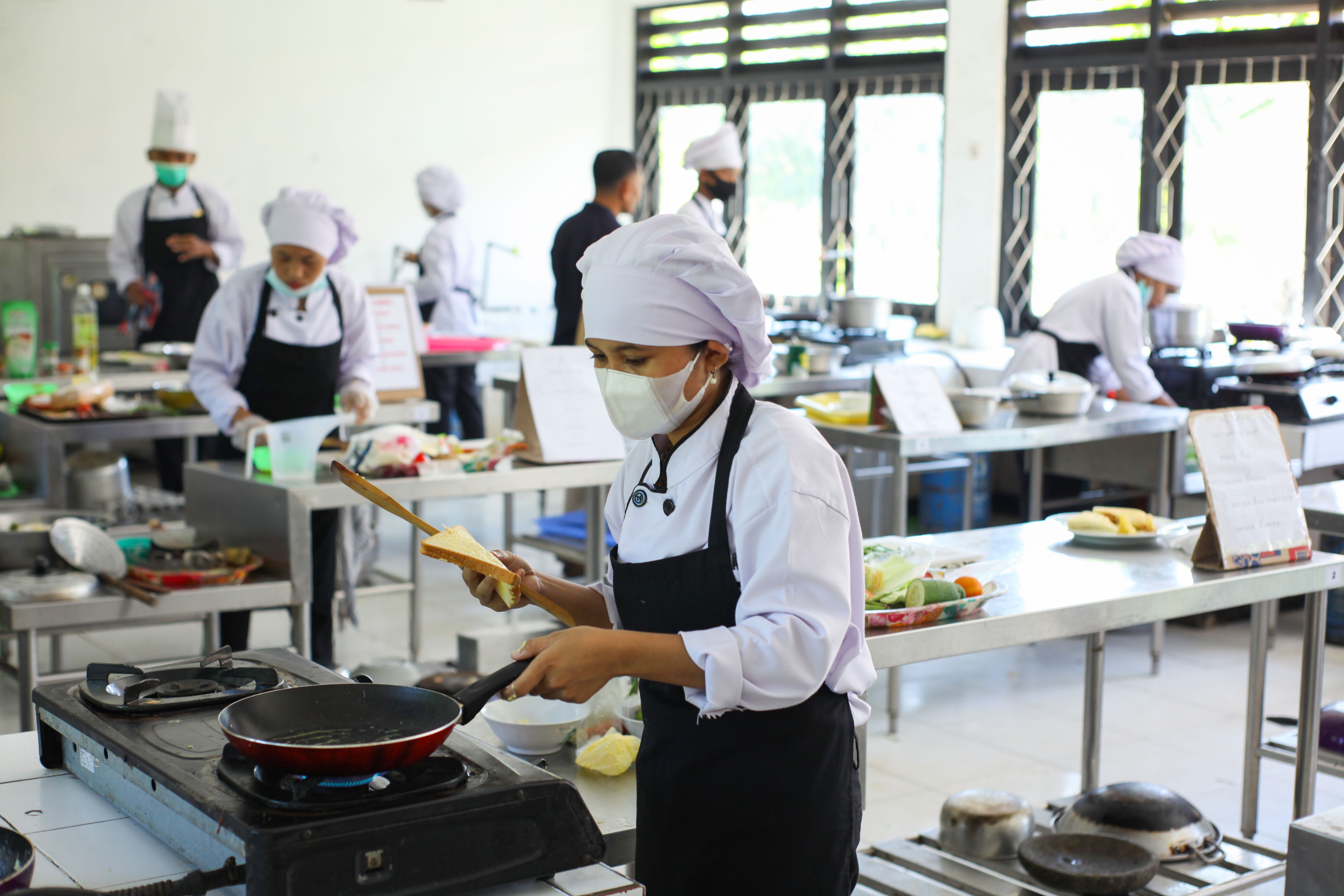 A female culinary student wearing a face mask and white chef uniform placing a slice of bread into a frying pan in a training kitchen.