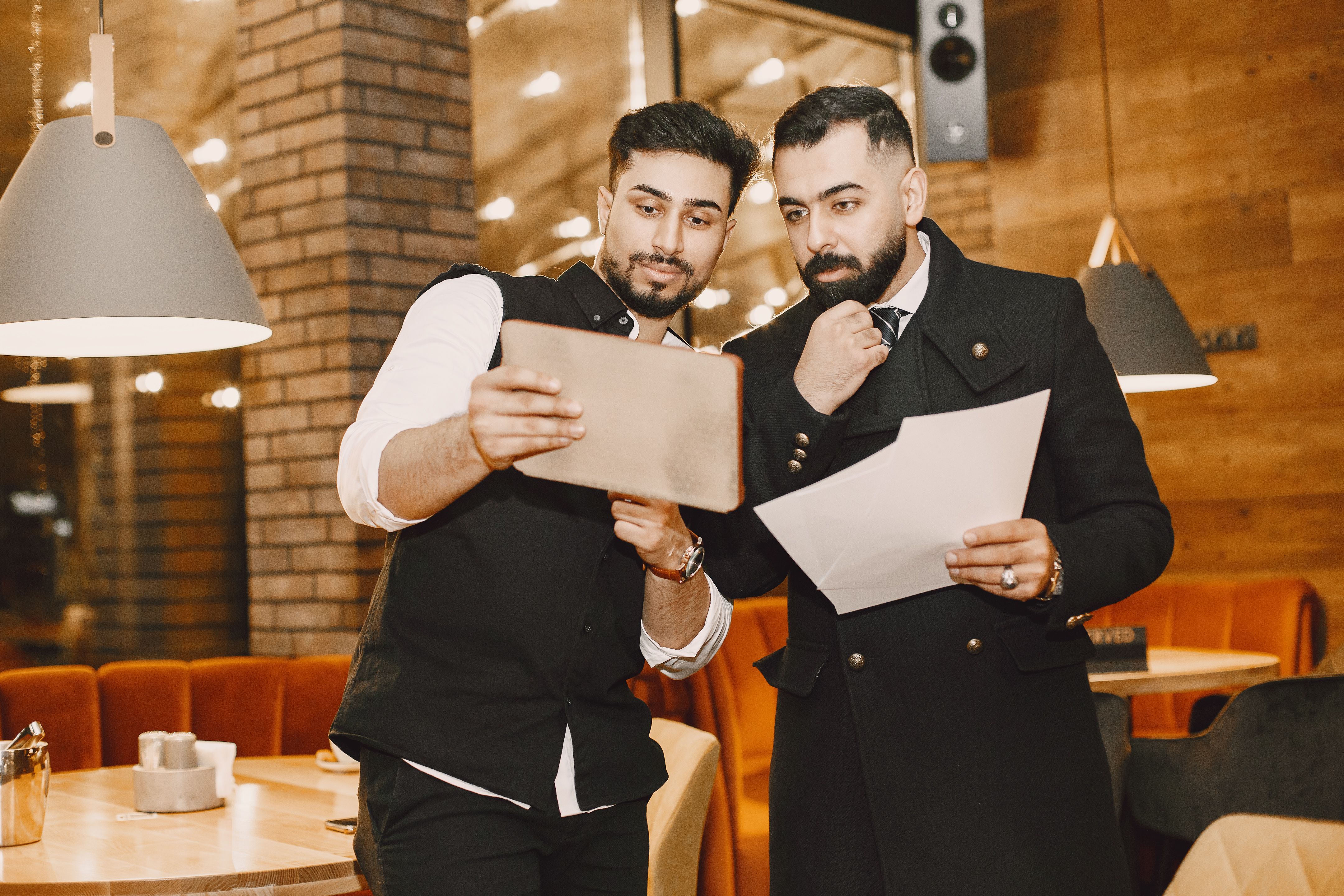 A restaurant manager showing information on a tablet to a colleague holding paper documents inside a dining area.