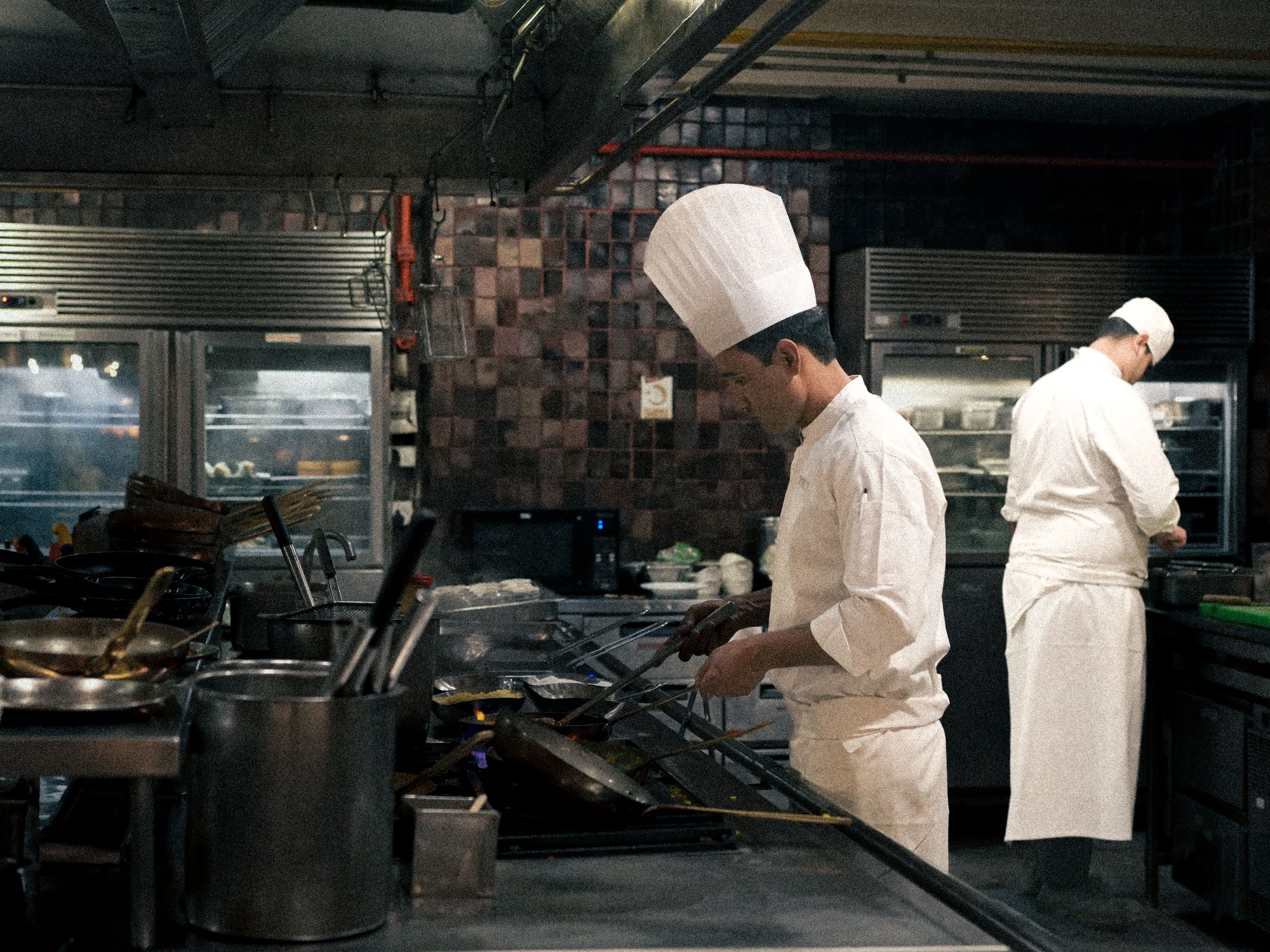 Two professional chefs wearing white uniforms and chef hats cooking food on stoves in a commercial restaurant kitchen.