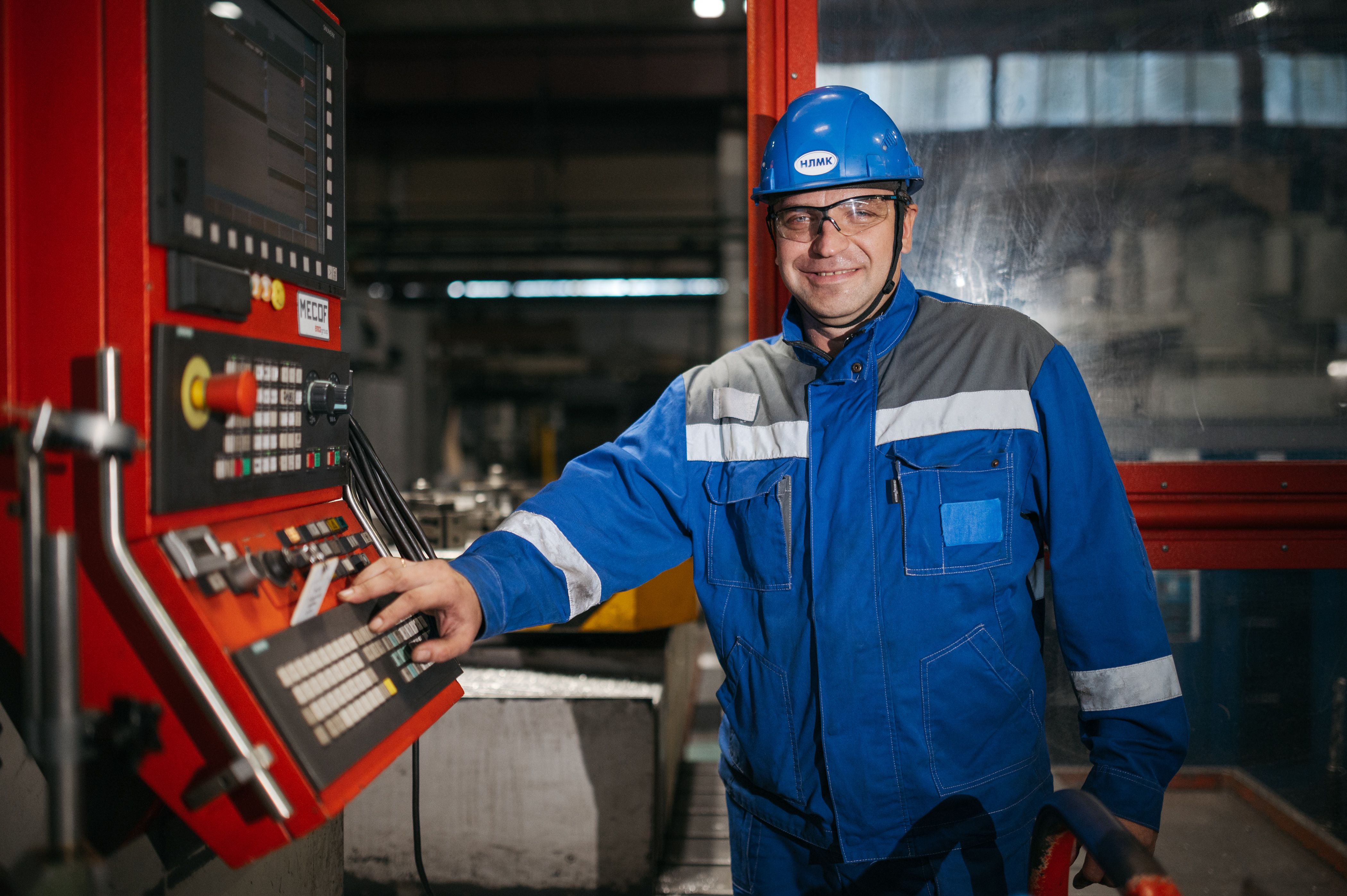 An industrial worker wearing a blue hard hat, protective glasses, and blue work overalls operating a machinery control panel.