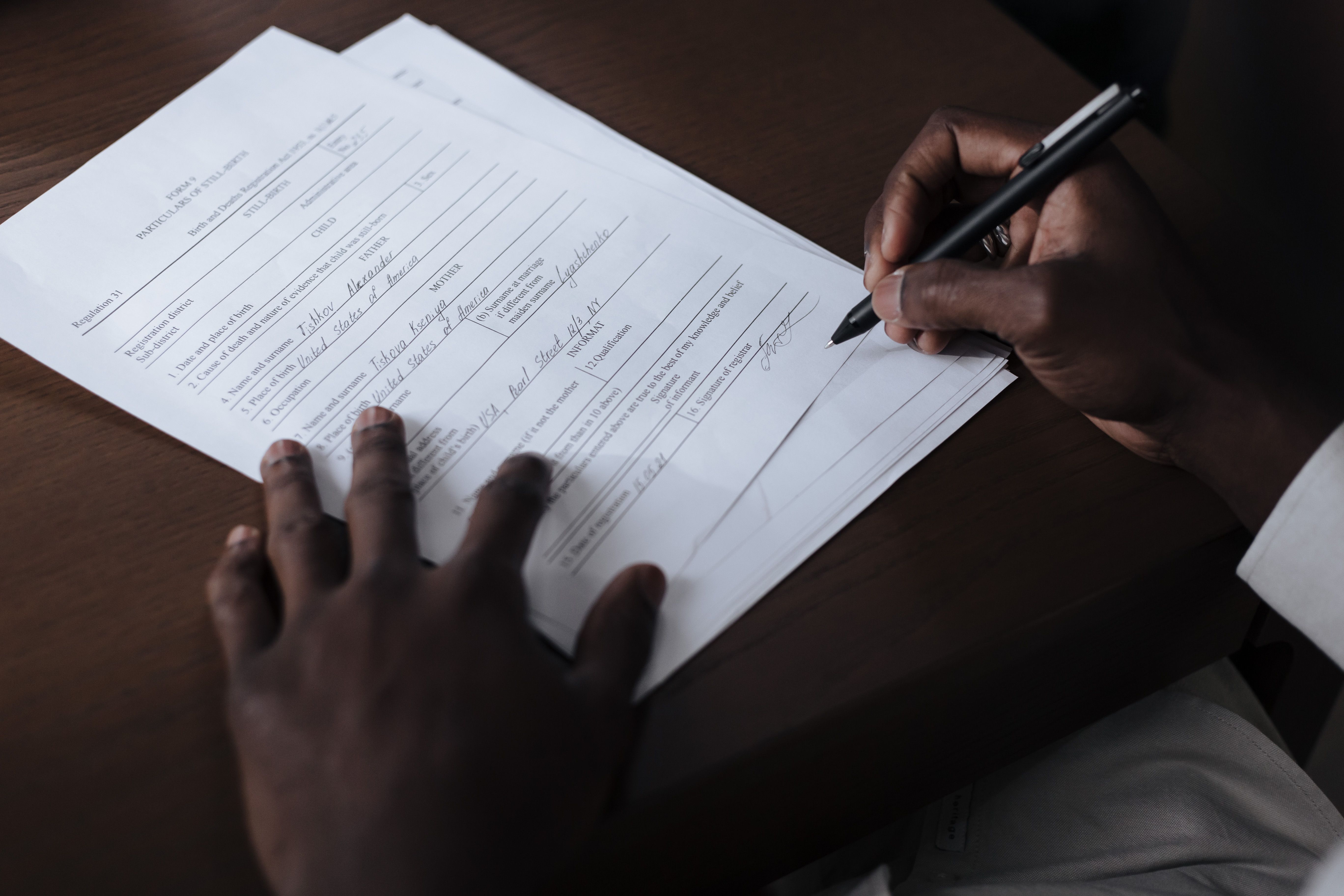 Overhead view of a person using a black pen to fill out official paper documents on a dark wooden desk.