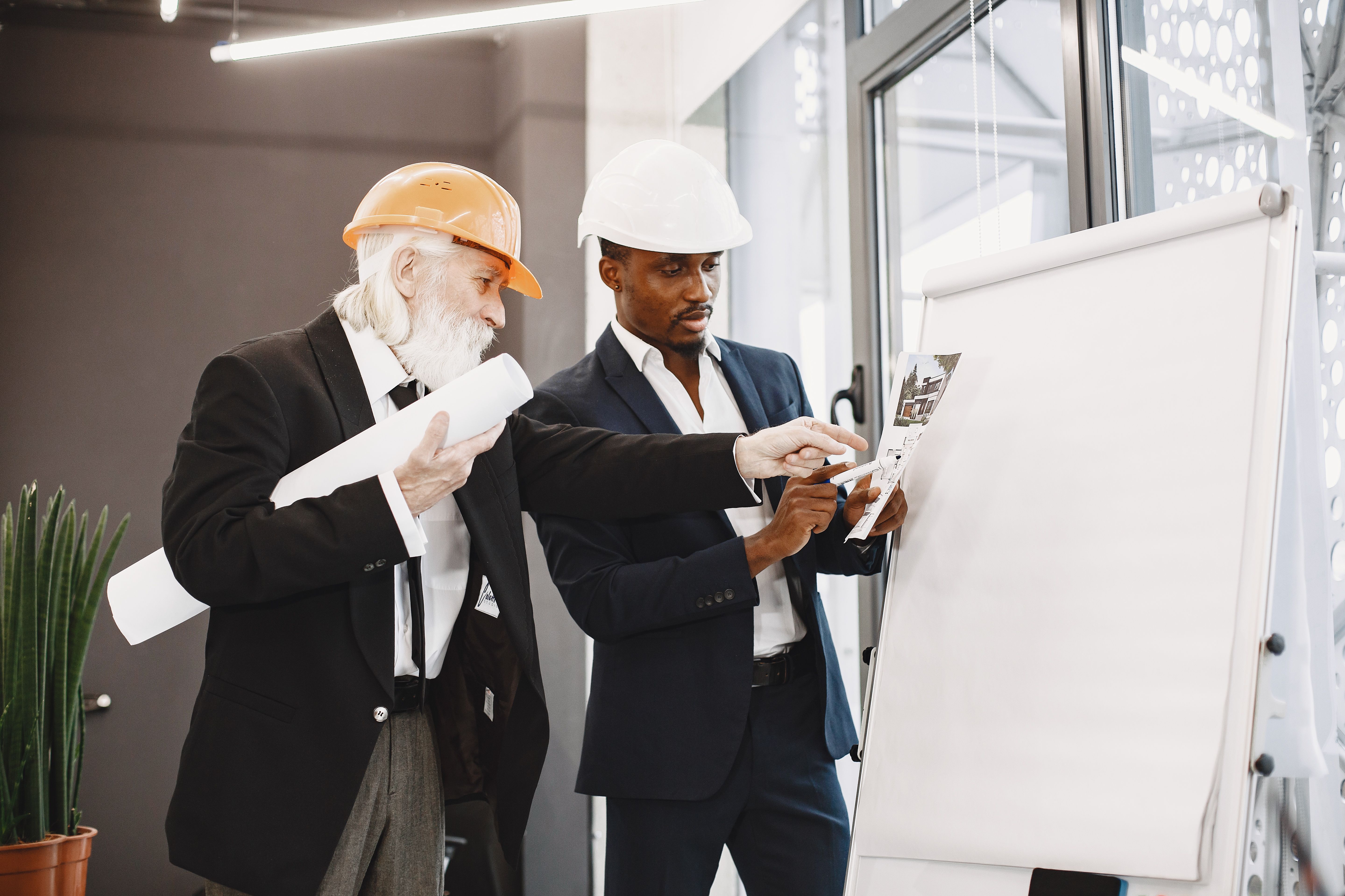 An older architect holding rolled blueprints discussing a project diagram with a young colleague in a suit near a whiteboard, both wearing hard hats.