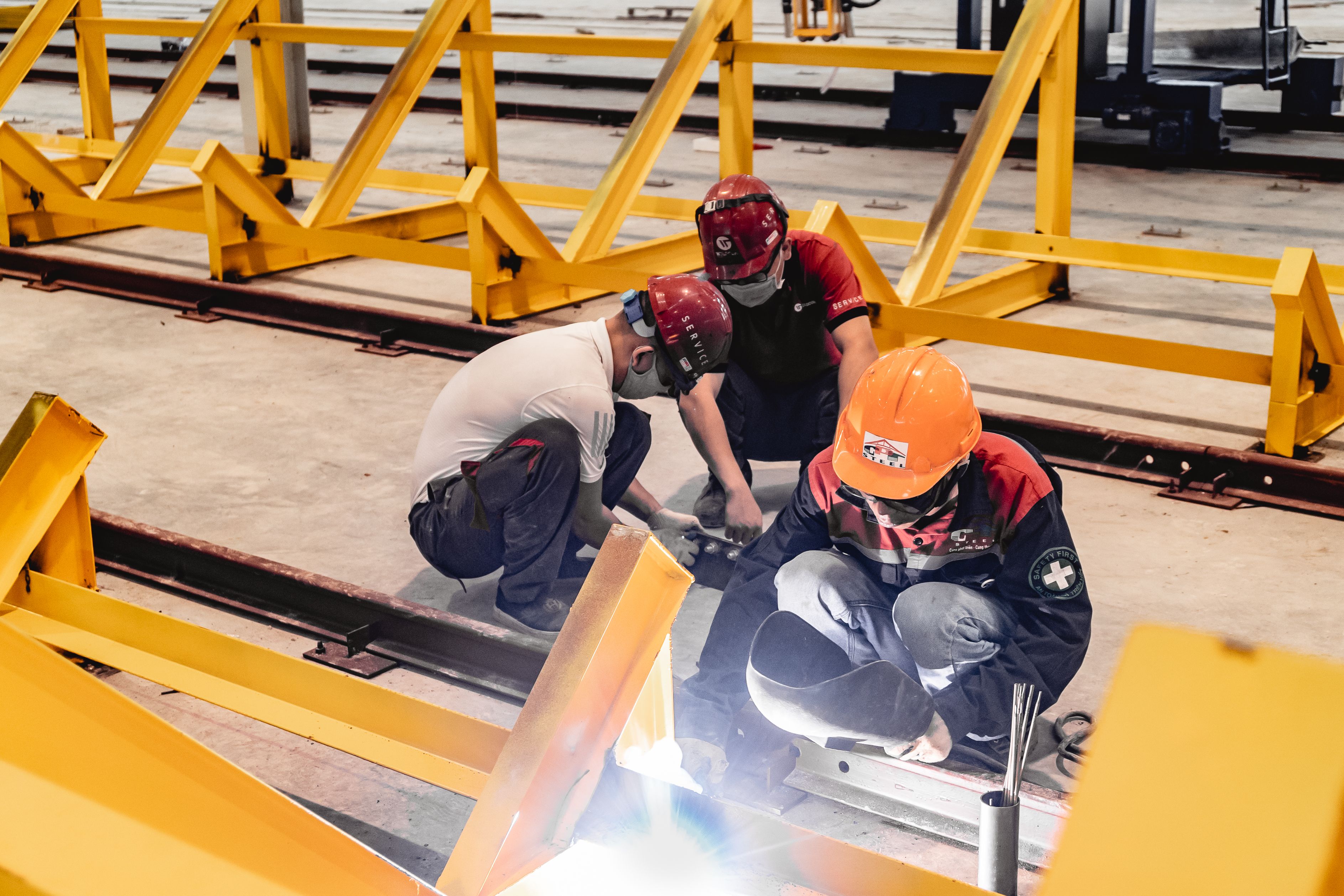 Three construction workers wearing hard hats and protective gear crouched down performing welding and assembly work on steel beams.