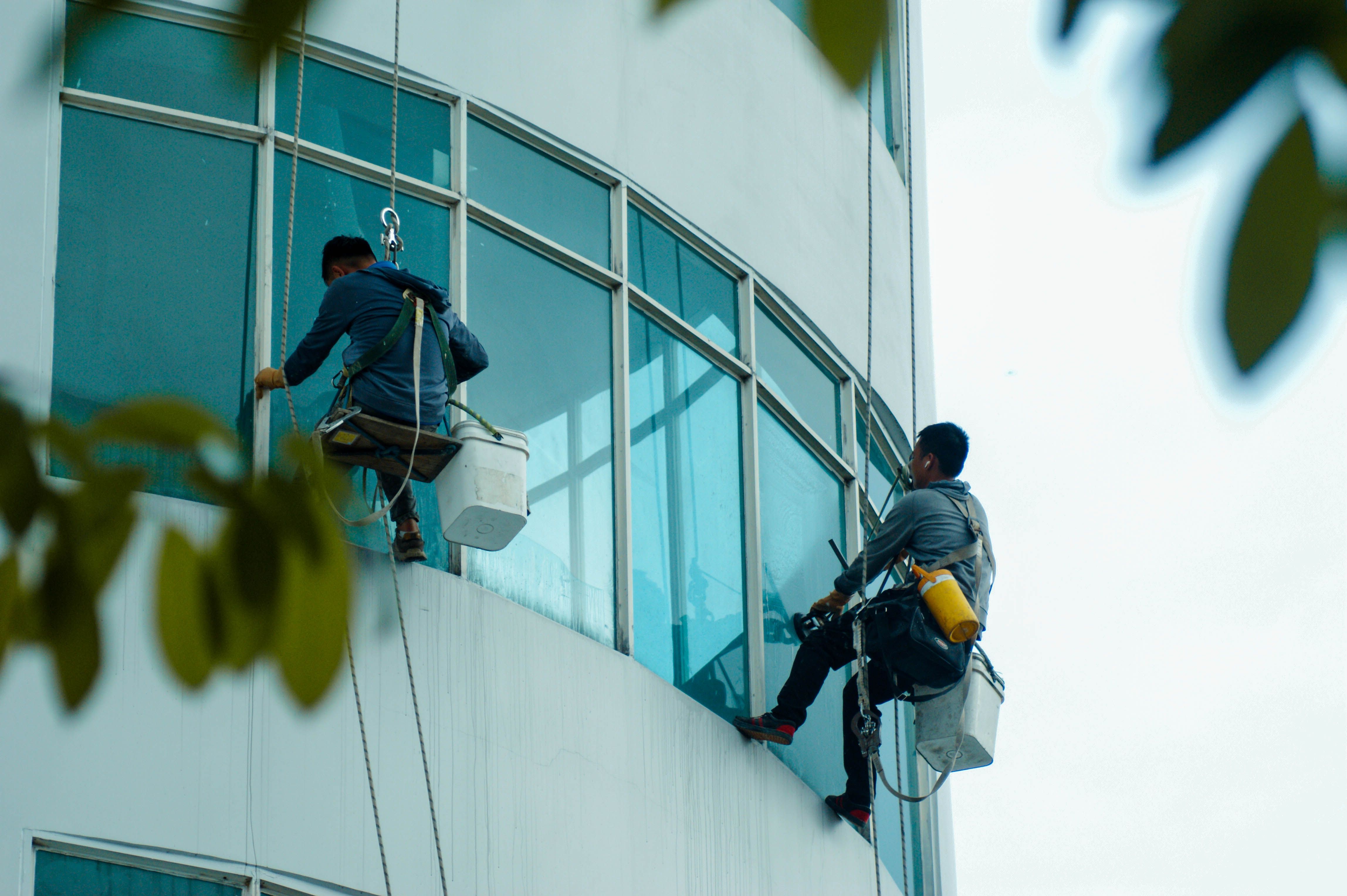 Two window cleaners wearing safety harnesses suspended from ropes cleaning the glass exterior of a tall building.