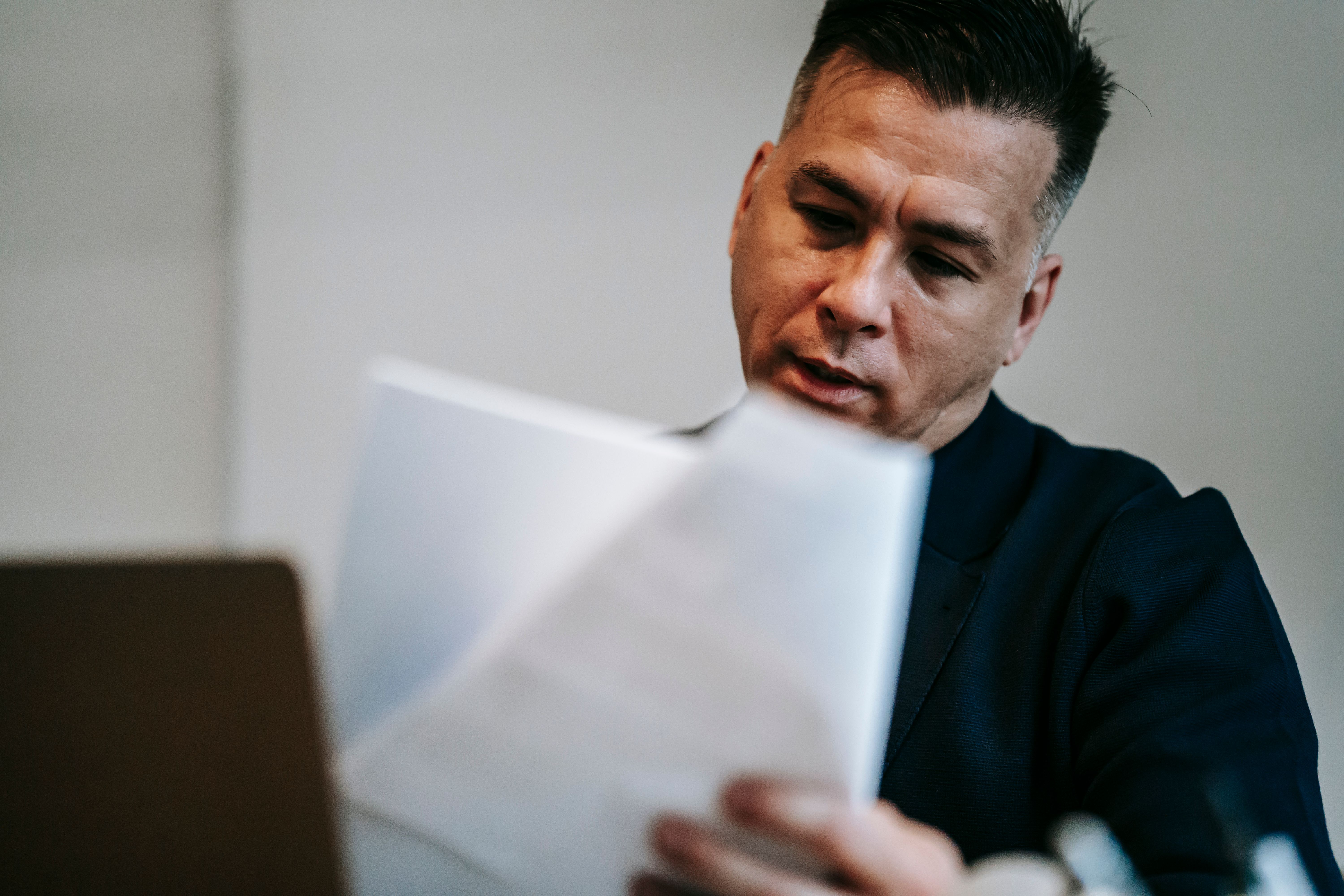 A businessman wearing a dark suit thoughtfully reviewing paper documents at his desk.