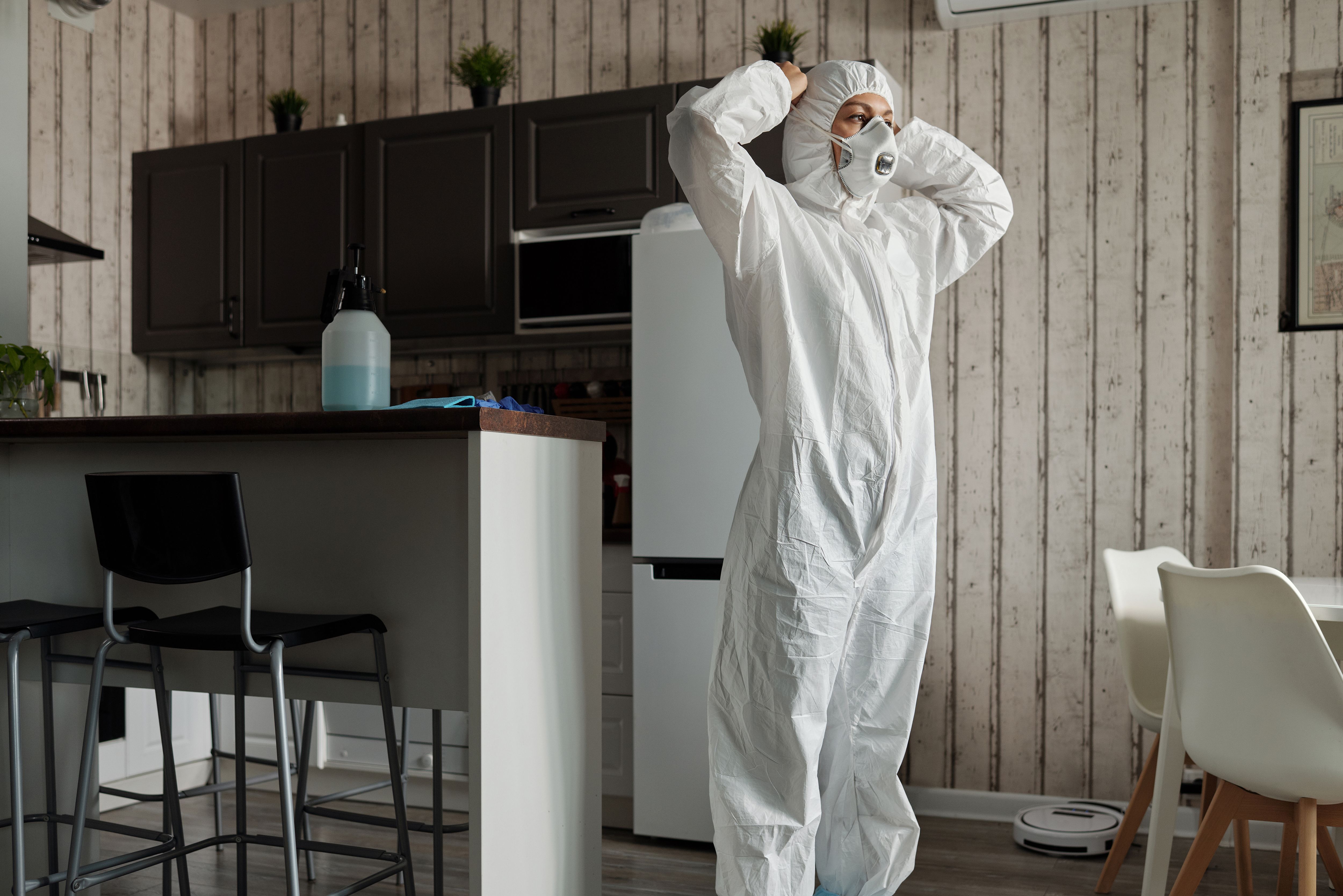 A pest control technician in a full-body white protective suit adjusting their mask inside a kitchen.