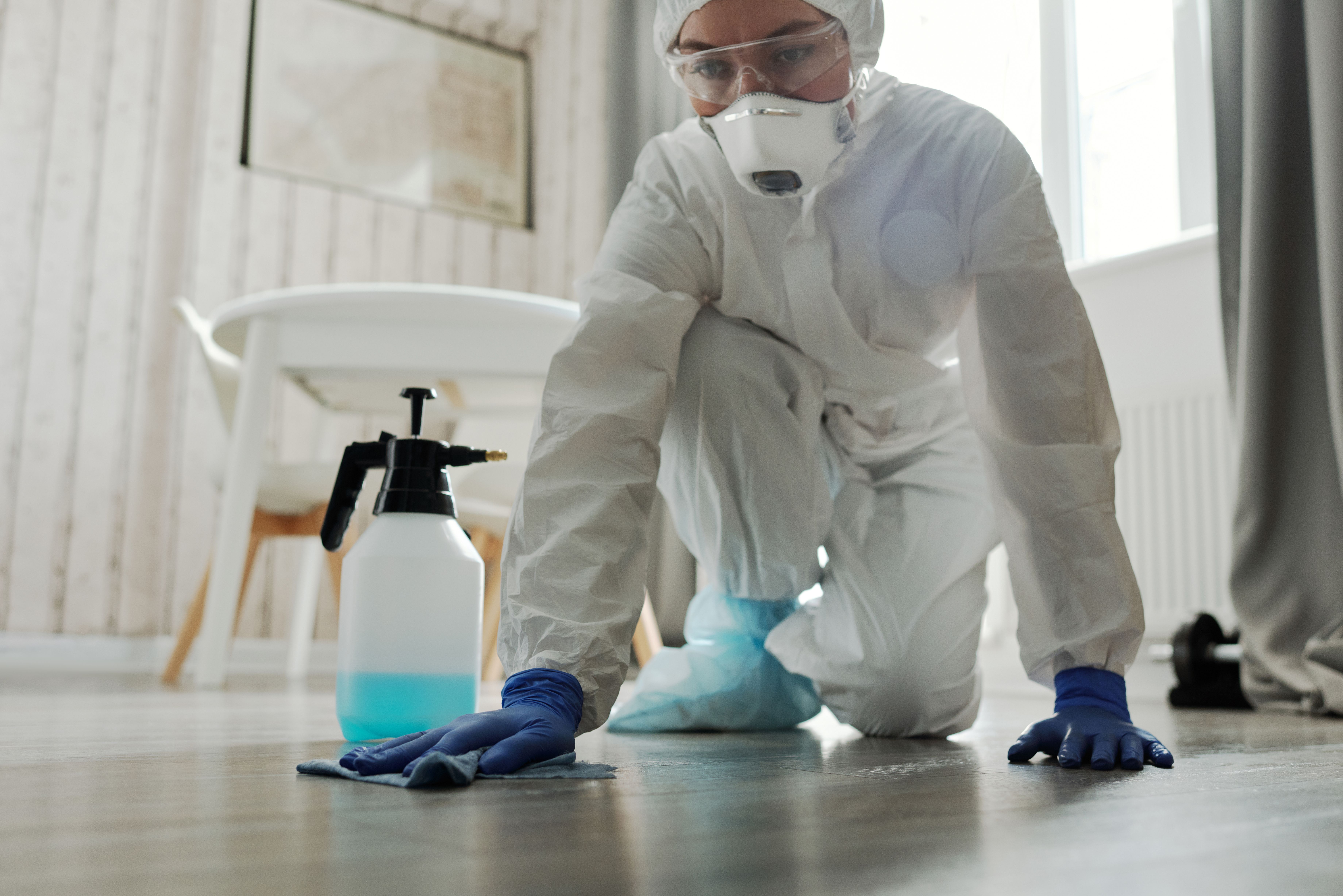 A person wearing a full protective hazmat suit and respirator mask kneeling to wipe a floor next to a chemical spray bottle.