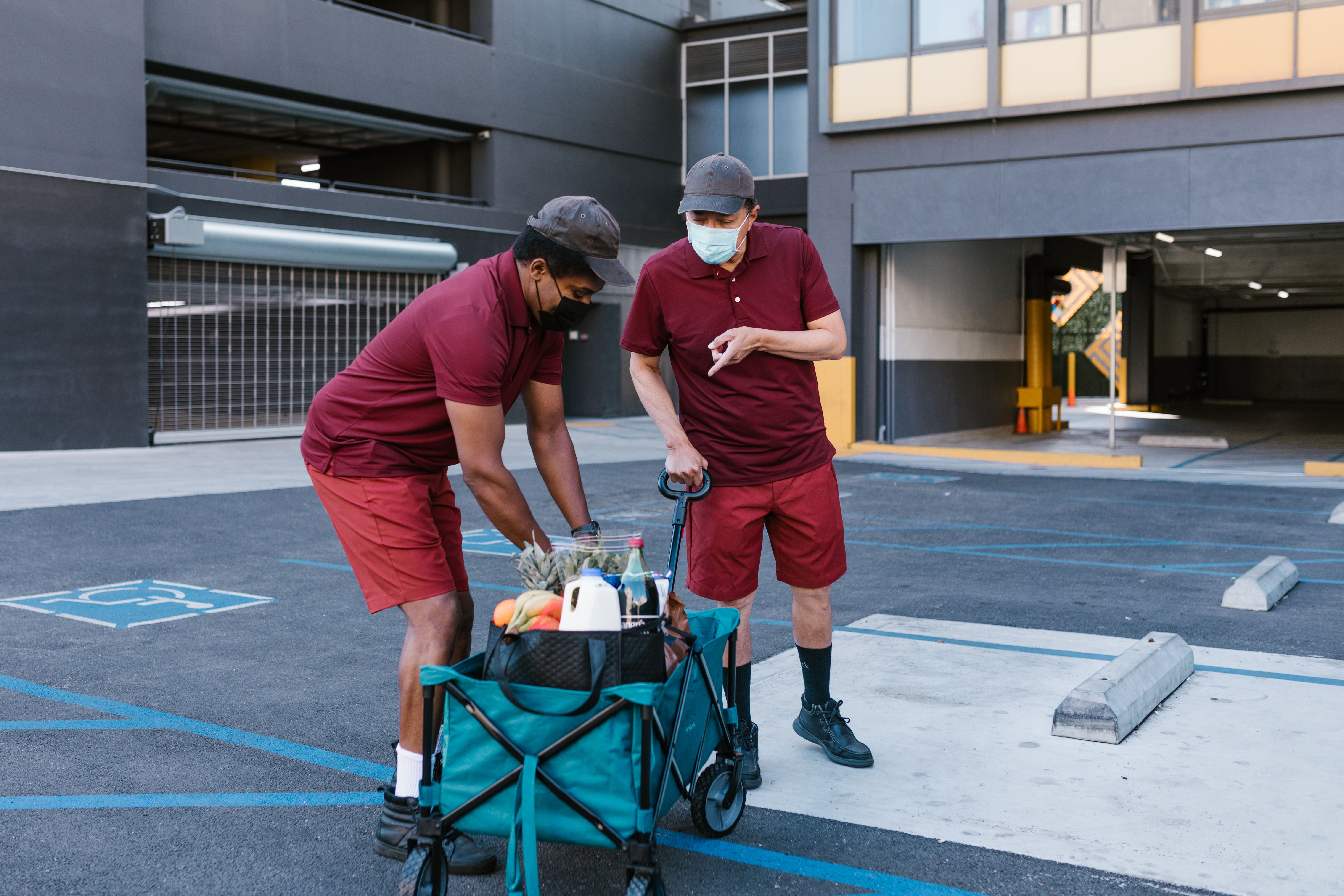 Two delivery workers wearing face masks and matching maroon uniforms loading fresh groceries into a utility cart in a parking structure.